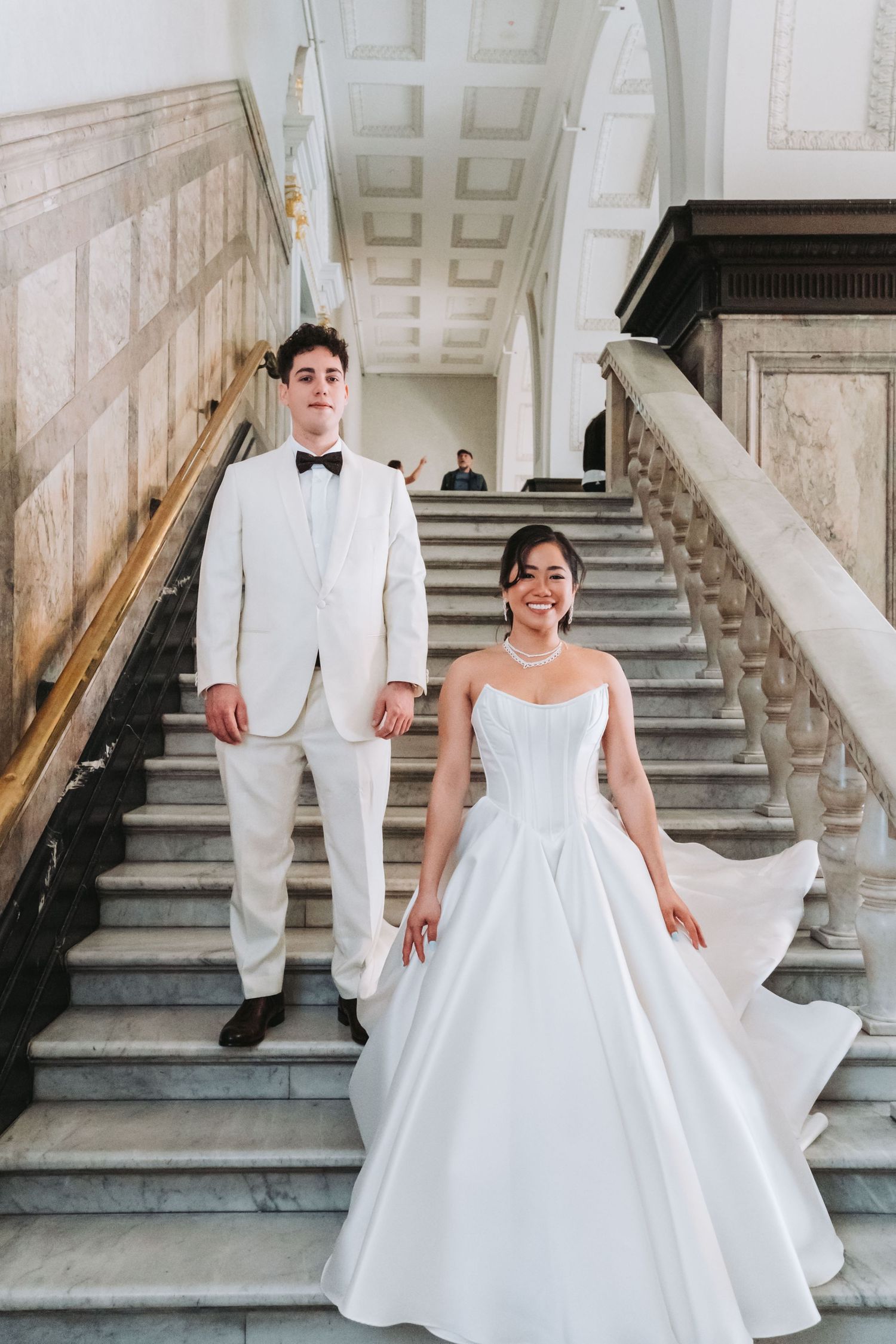 Bride in white ballgown and groom in ivory tuxedo pose on elegant marble staircase for wedding portrait.