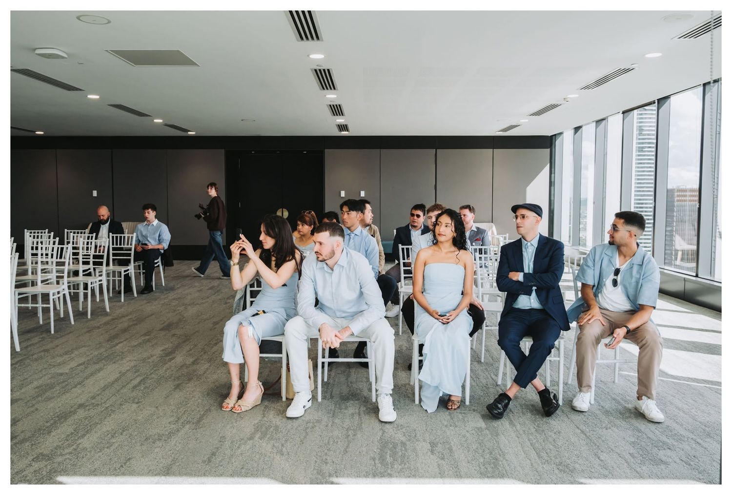 Guests seated in white chairs at an elegant indoor wedding ceremony with floor-to-ceiling windows.