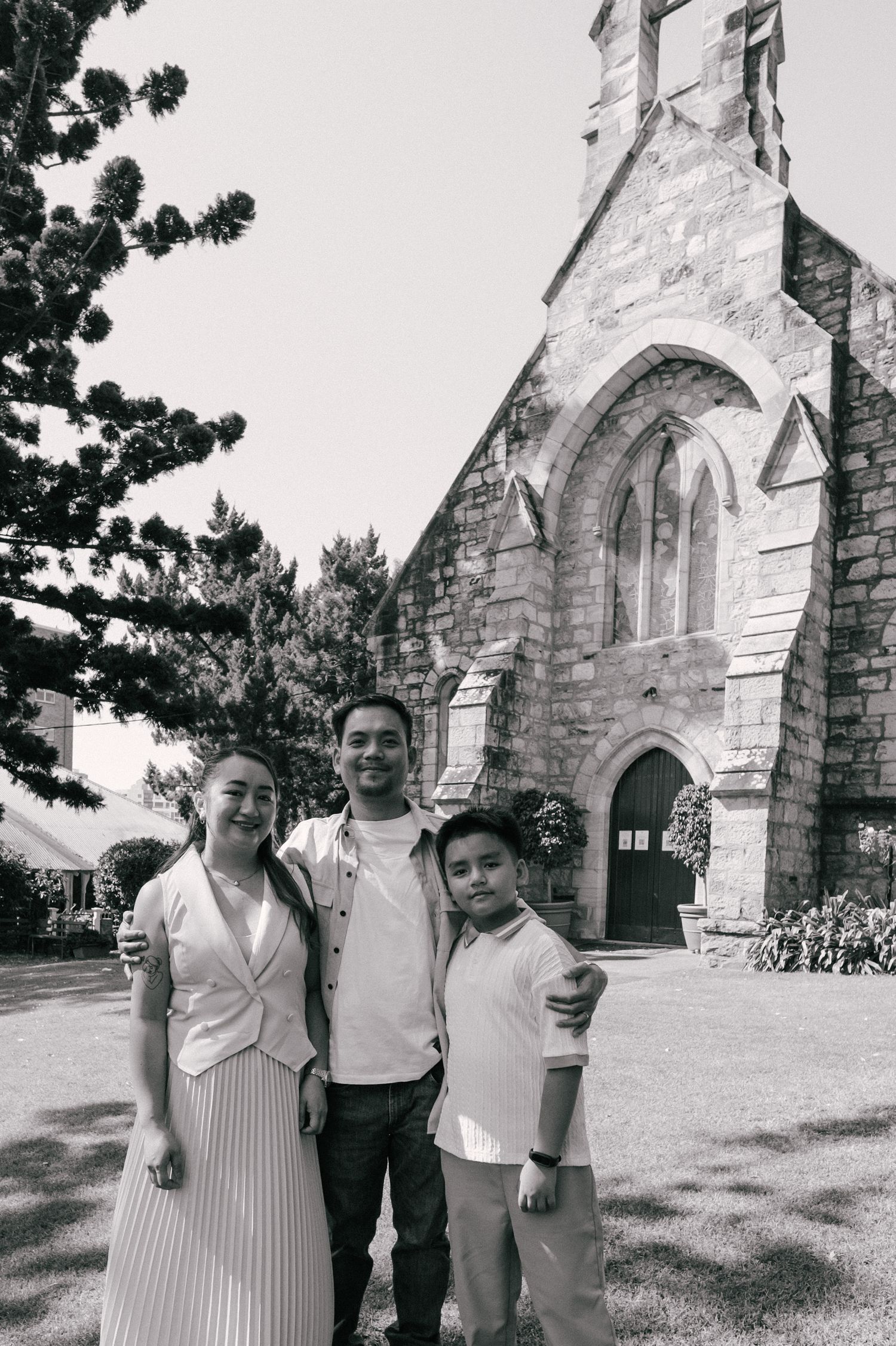 Black and white photo of a smiling family of three posing outside a historic stone church on a sunny day.