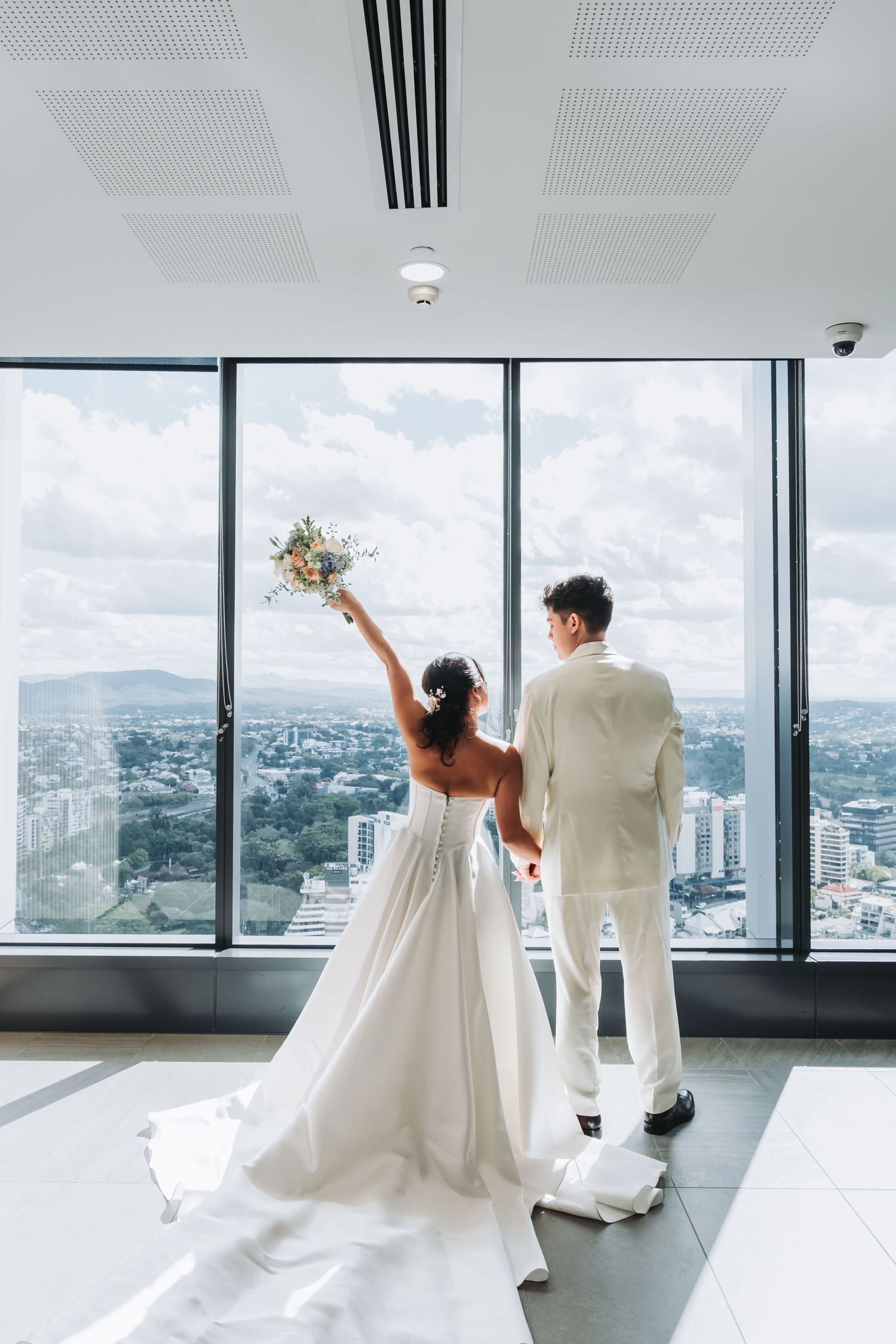 Bride raising bouquet with groom before panoramic city view windows in a high-rise wedding venue.