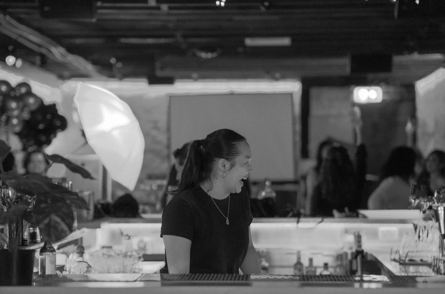 Black and white photo of a woman working behind a bar counter with a large white umbrella light in the background.