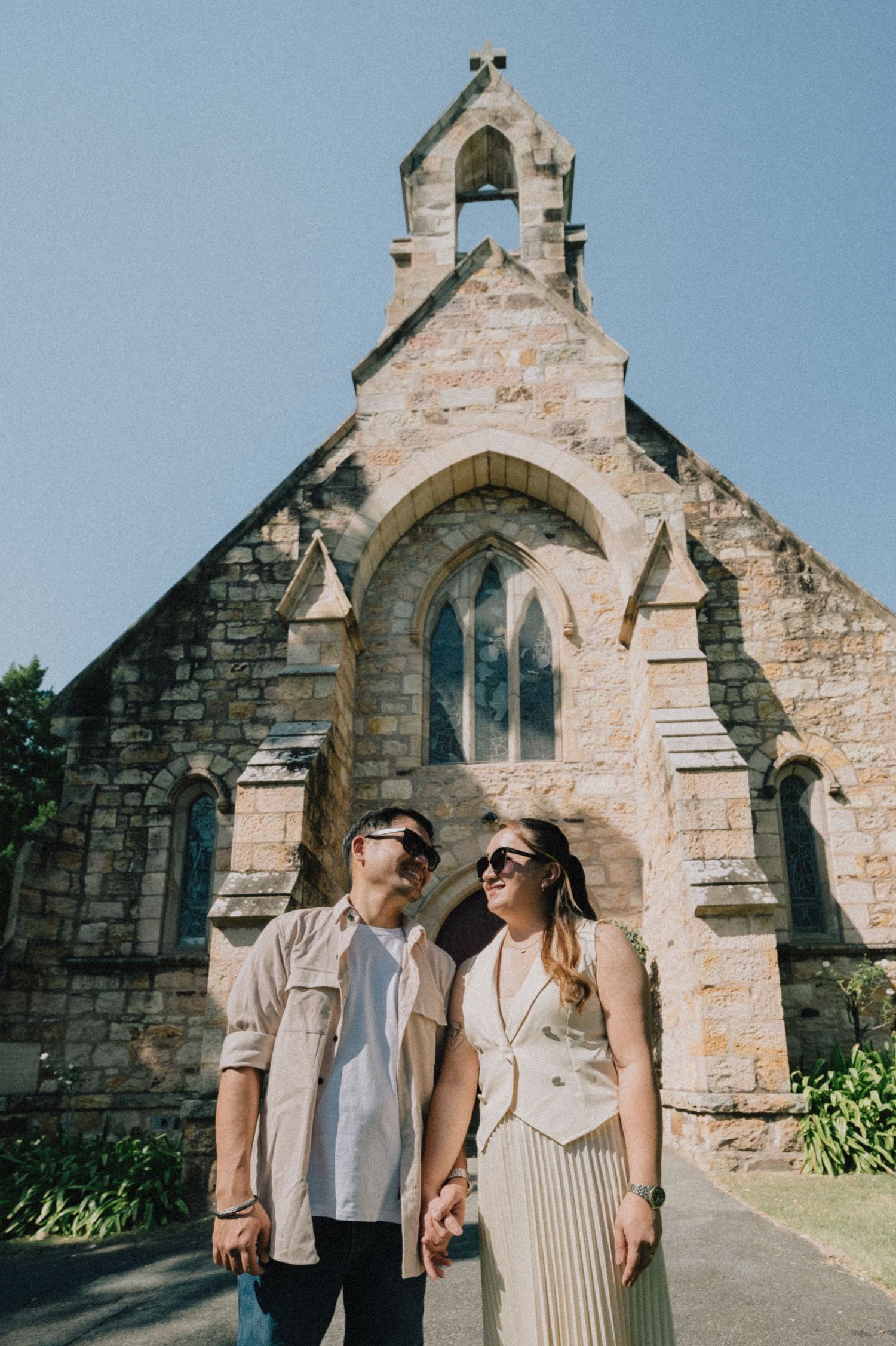 Couple holding hands in front of a historic stone church on a sunny day, wearing casual outfits and sunglasses.