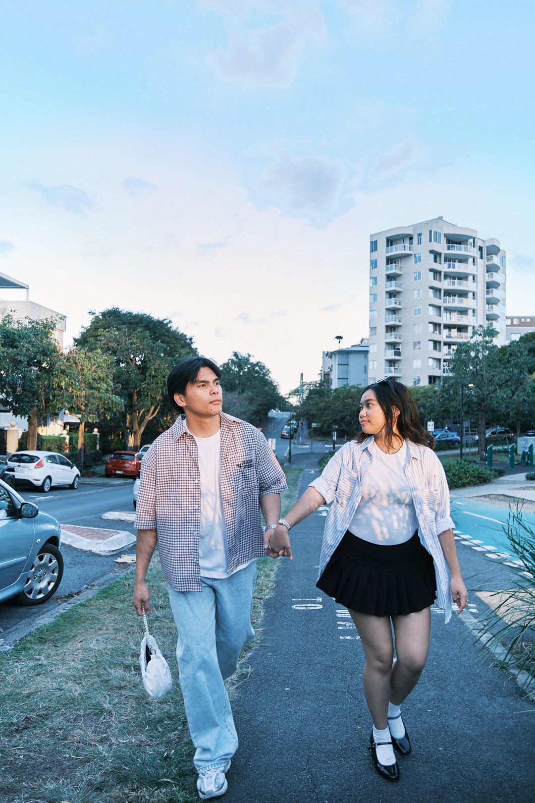 Couple holding hands walking on urban sidewalk, woman in black skirt and man in jeans, city buildings in background.