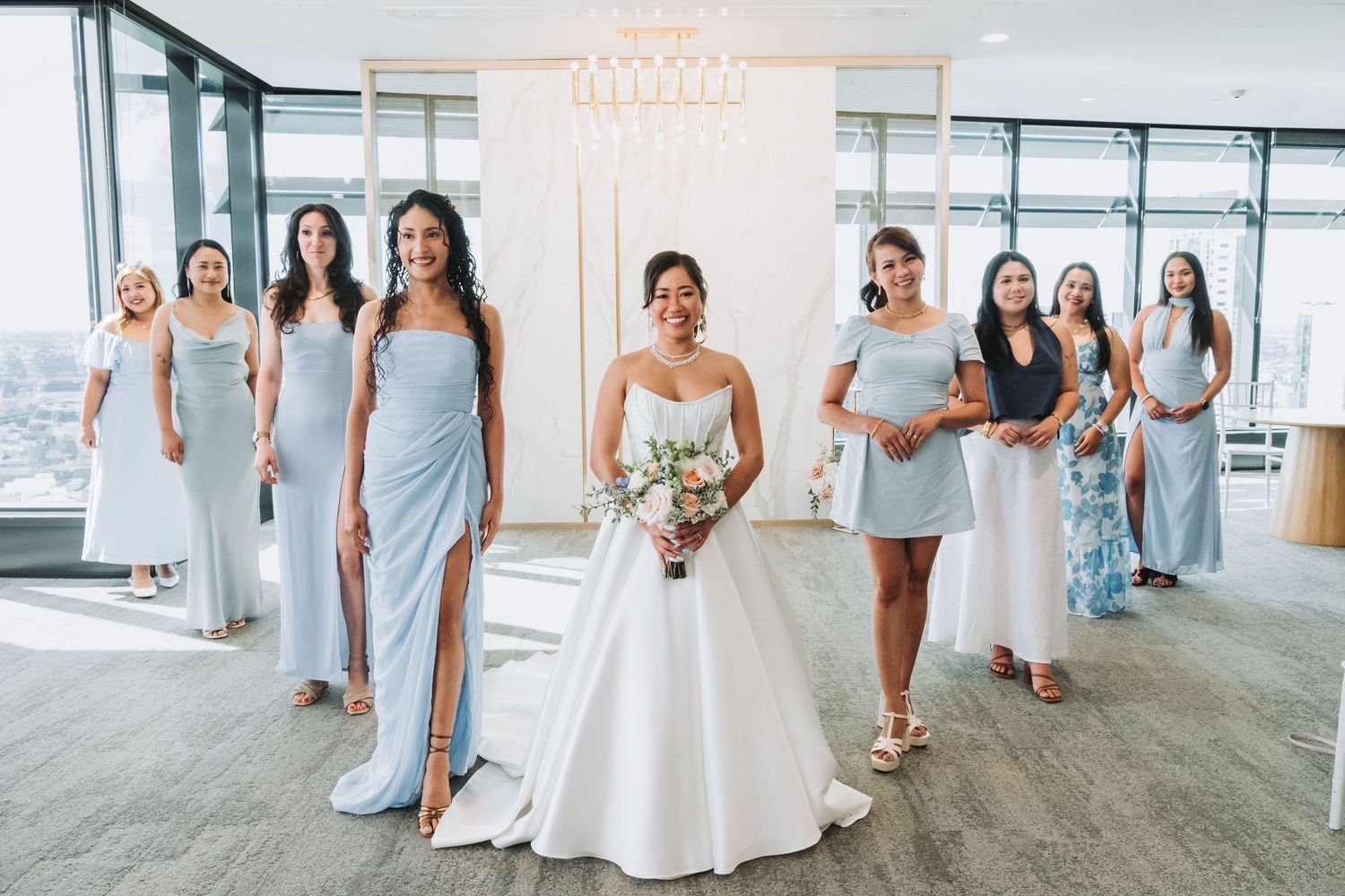 Bride in white ball gown holds bouquet surrounded by bridesmaids in light blue dresses in elegant venue.