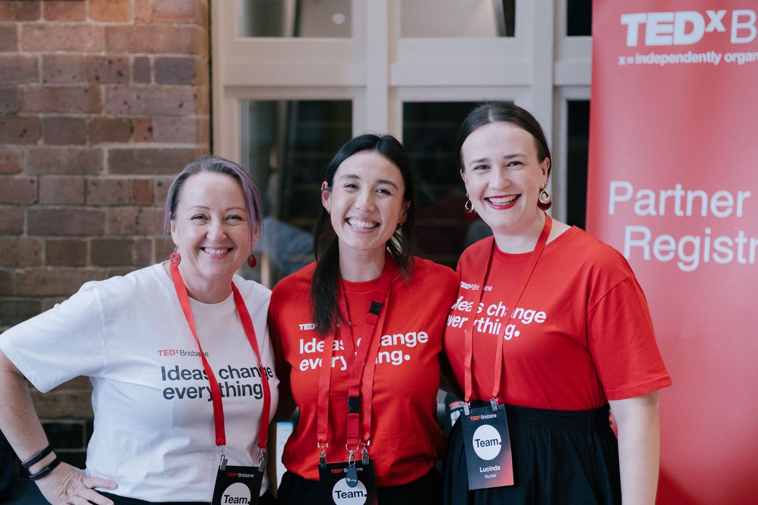 Three women in red TEDx Ideas Change Everything t-shirts smile together at a TEDx event.