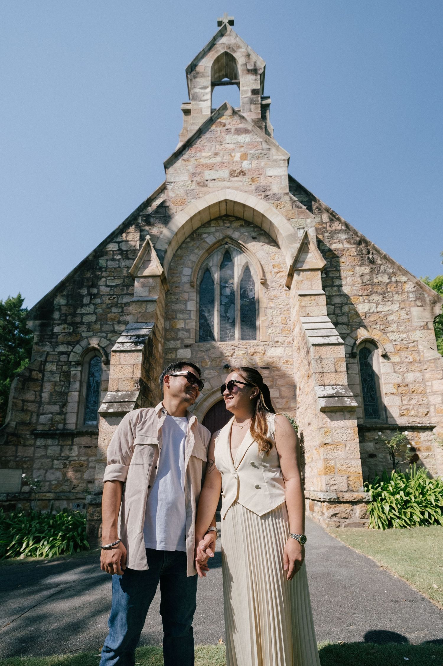 Couple holding hands in front of a historic stone church with Gothic architecture on a sunny day.