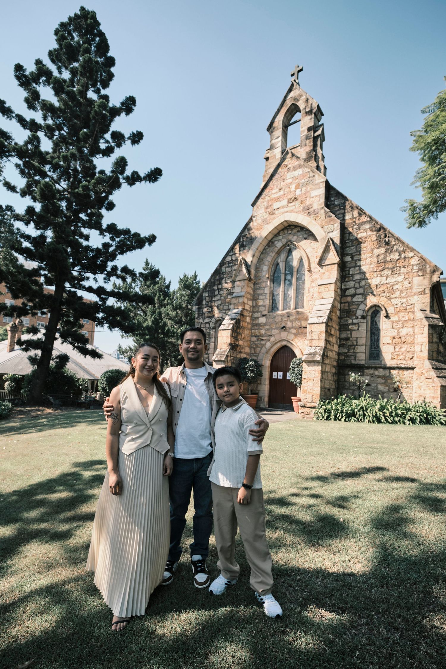 A family of three standing and posing in front of a stone church building.