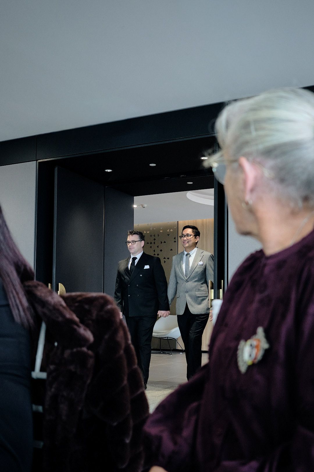 Two grooms in suits hold hands entering a room as guests in burgundy fur coats look on at a wedding.