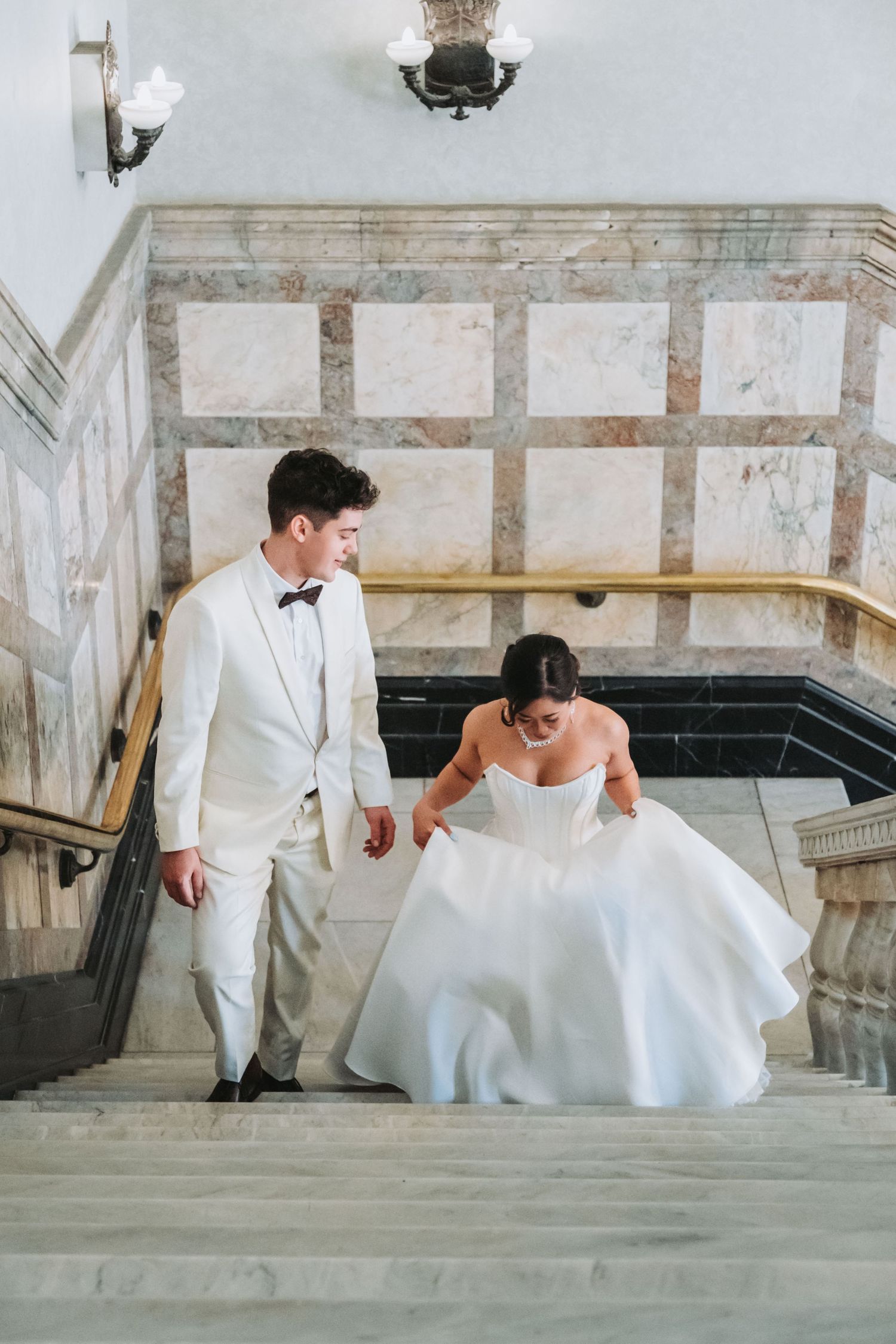 Bride in white gown and groom in ivory tuxedo walk up marble staircase in elegant wedding venue.