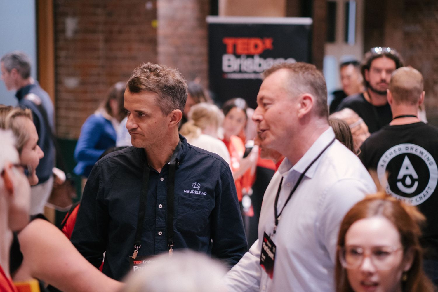 Two men networking at TEDx Brisbane event, engaged in conversation among a crowd of attendees indoors.