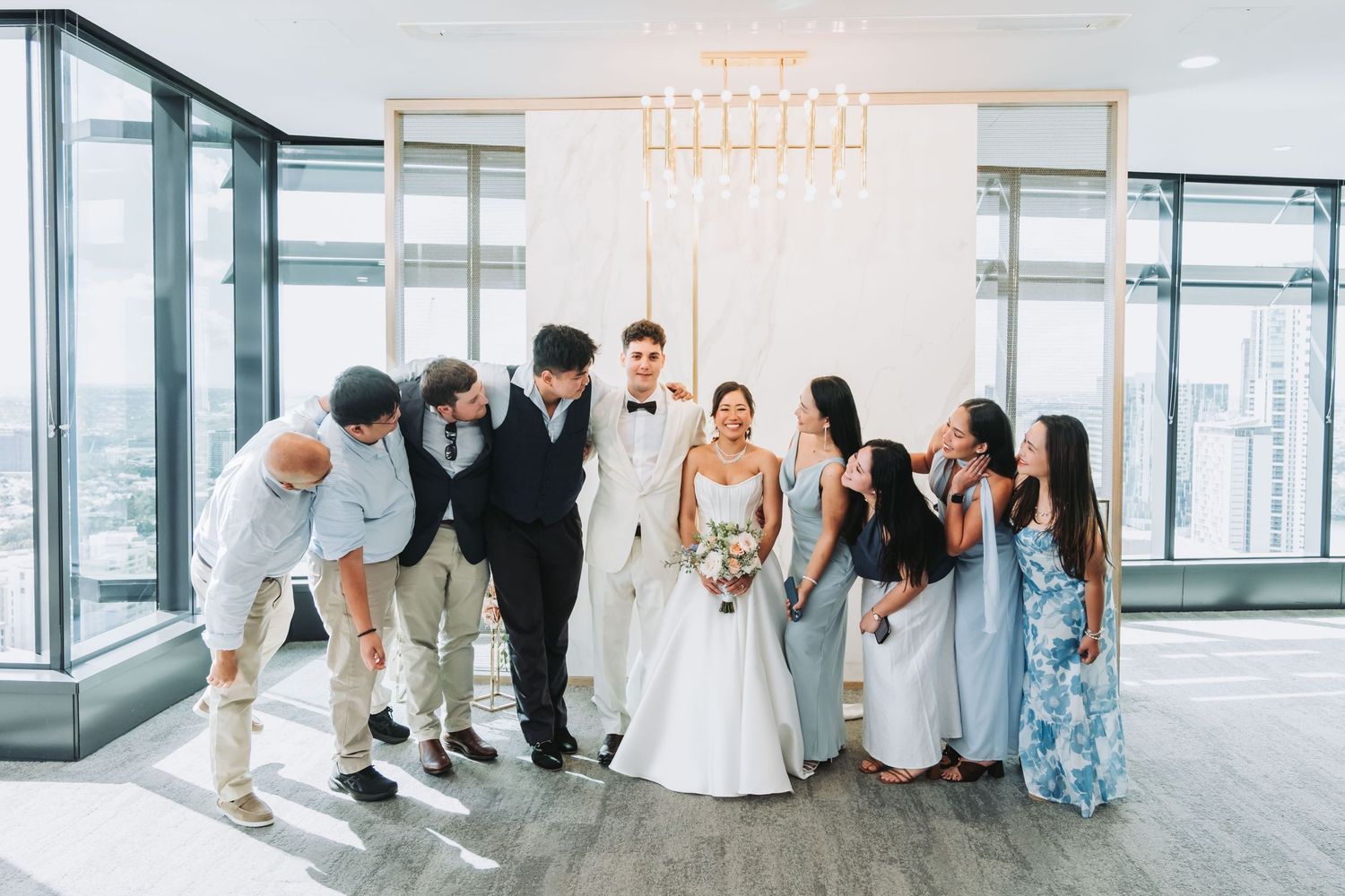 Wedding party celebrating with bride and groom in elegant high-rise venue with chandelier backdrop.