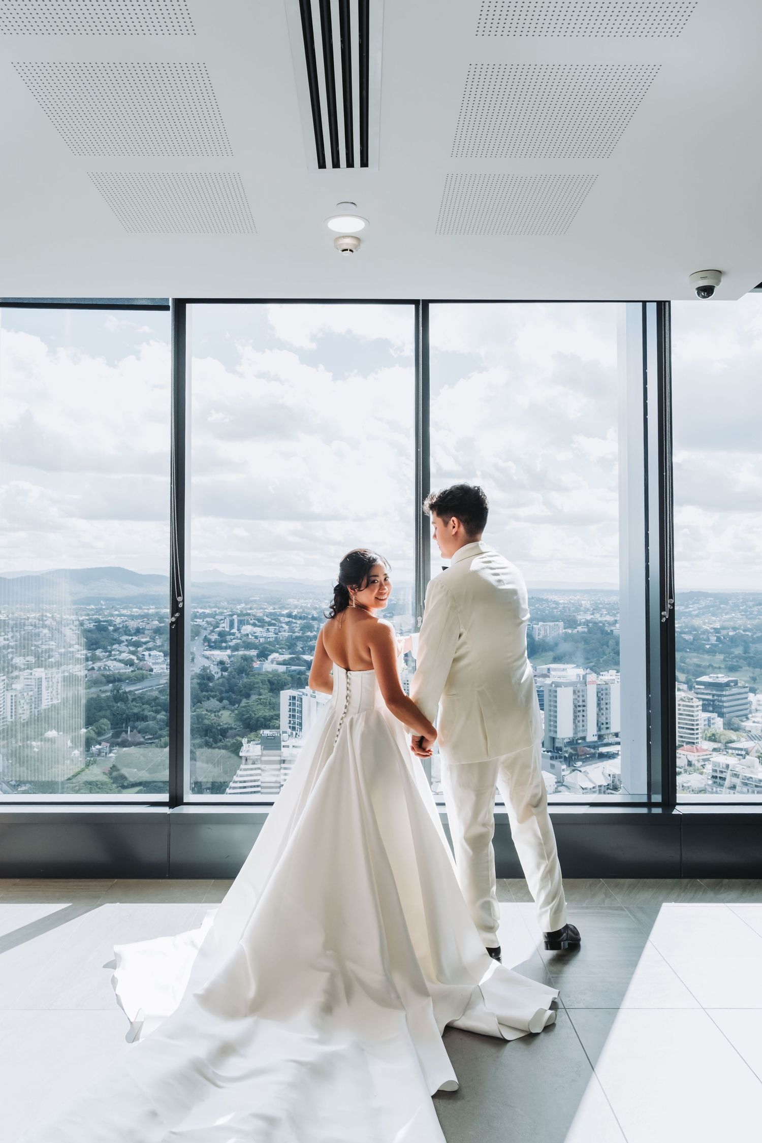Bride and groom holding hands in white wedding attire, standing before floor-to-ceiling windows with city skyline view.
