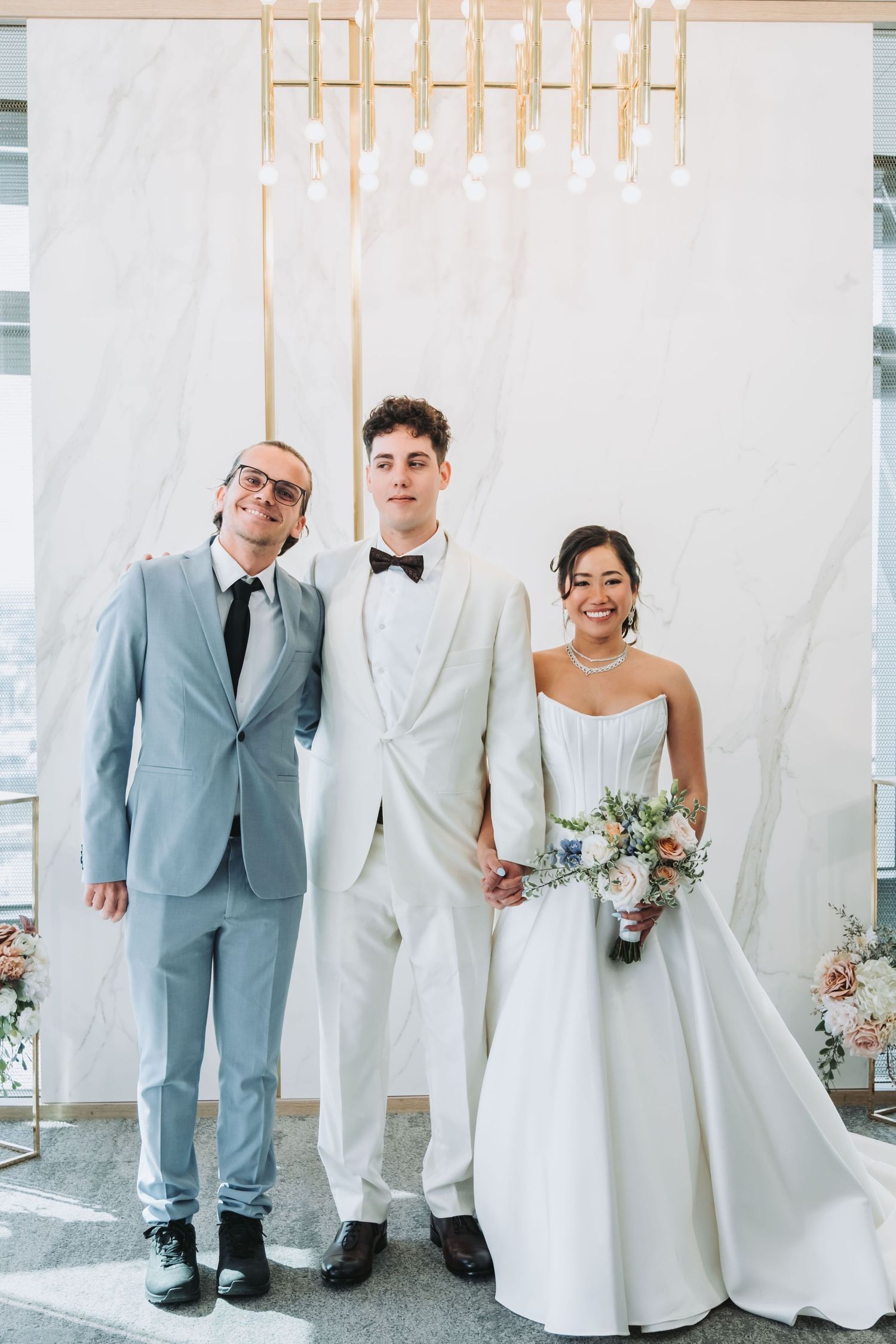 Bride in white gown holding bouquet stands with groom in white tuxedo and guest in gray suit at elegant wedding ceremony.
