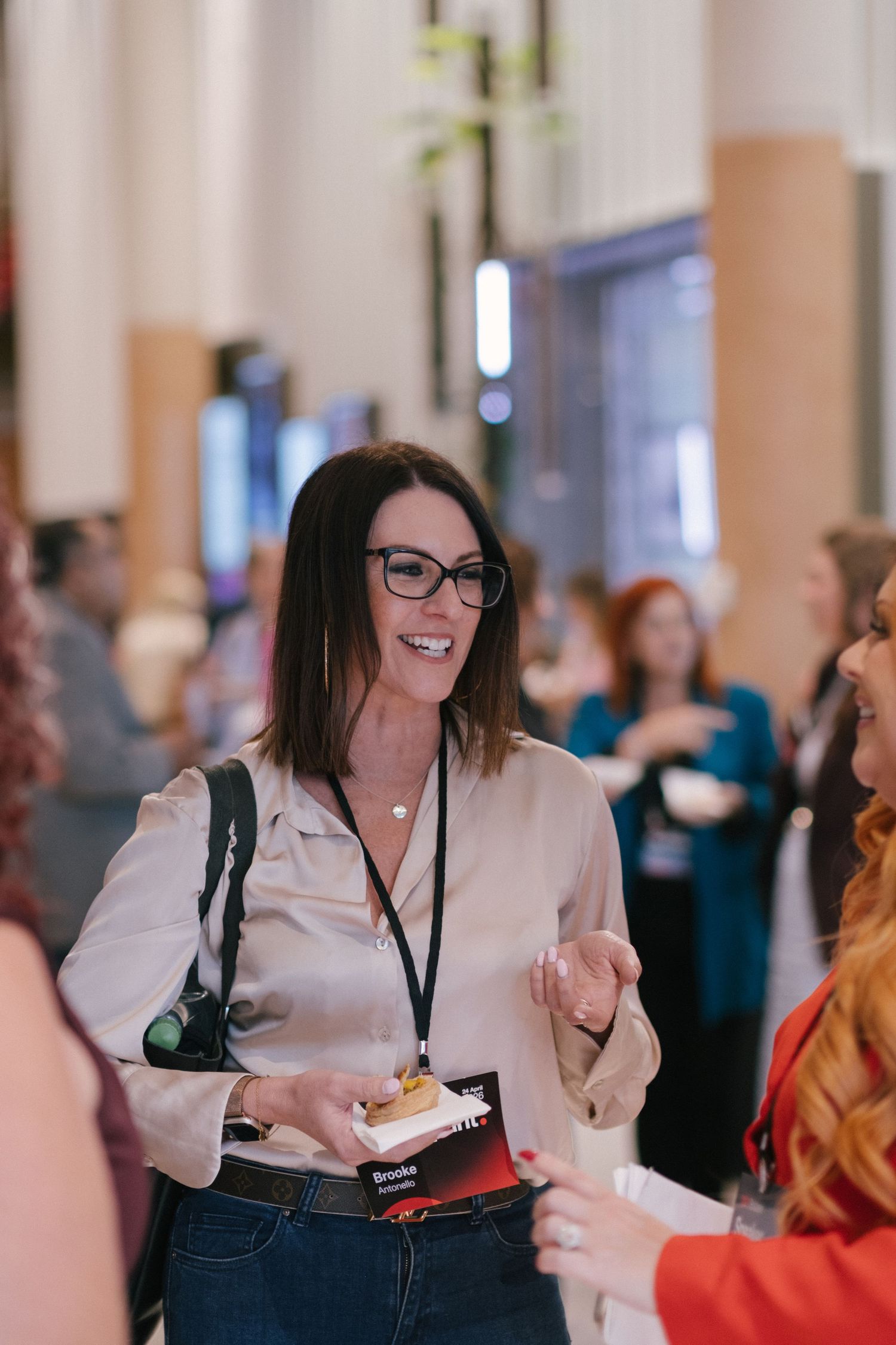 Woman with glasses and lanyard smiling at a professional networking event, holding materials while engaging in conversation.