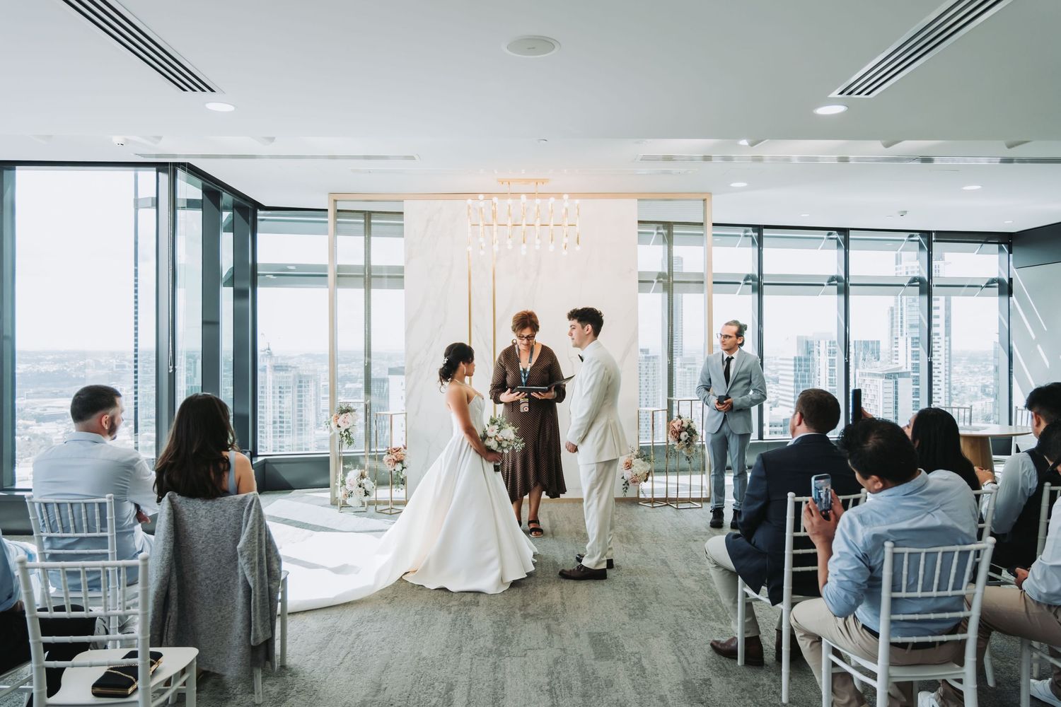 Elegant indoor wedding ceremony with bride and groom exchanging vows before officiant, floor-to-ceiling windows with city views.
