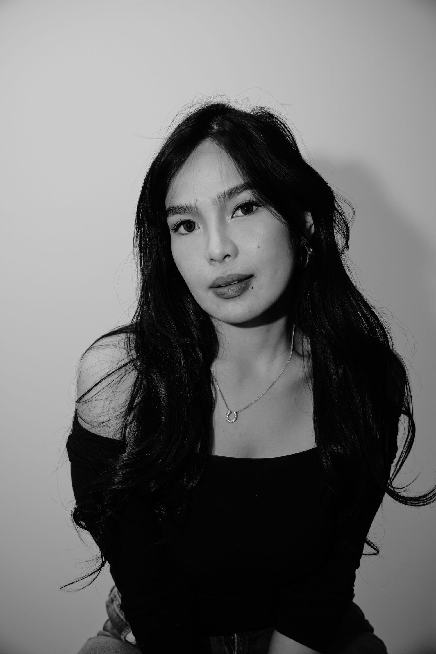 Black and white portrait of a young Asian woman with long dark hair wearing an off-shoulder black top and delicate necklace.