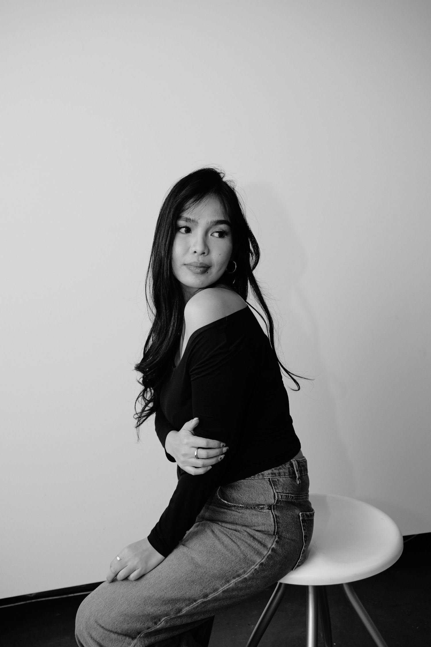 Black and white portrait of a young woman in off-shoulder top and jeans sitting on a stool against a white wall.