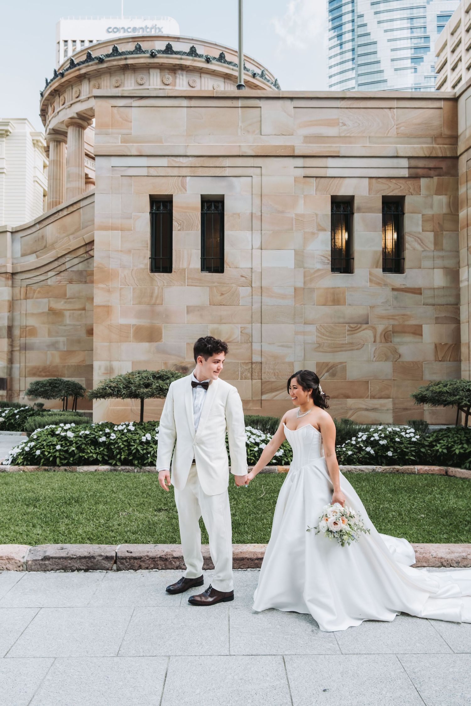 Bride and groom hold hands in front of a sandstone building in Brisbane, she in a white gown, he in a white suit.