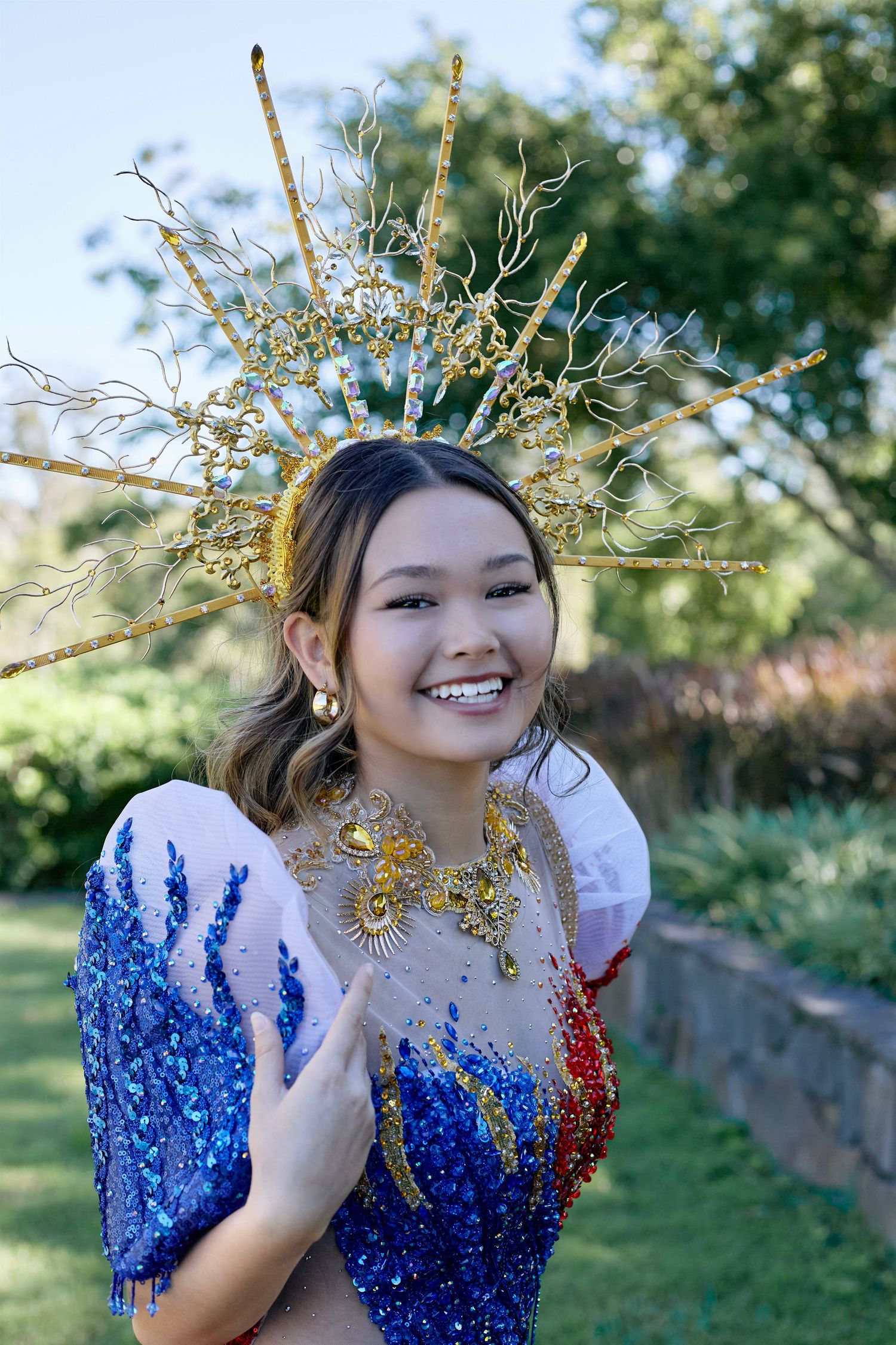 Young woman smiling in colorful sequined costume with ornate gold crown headdress outdoors.
