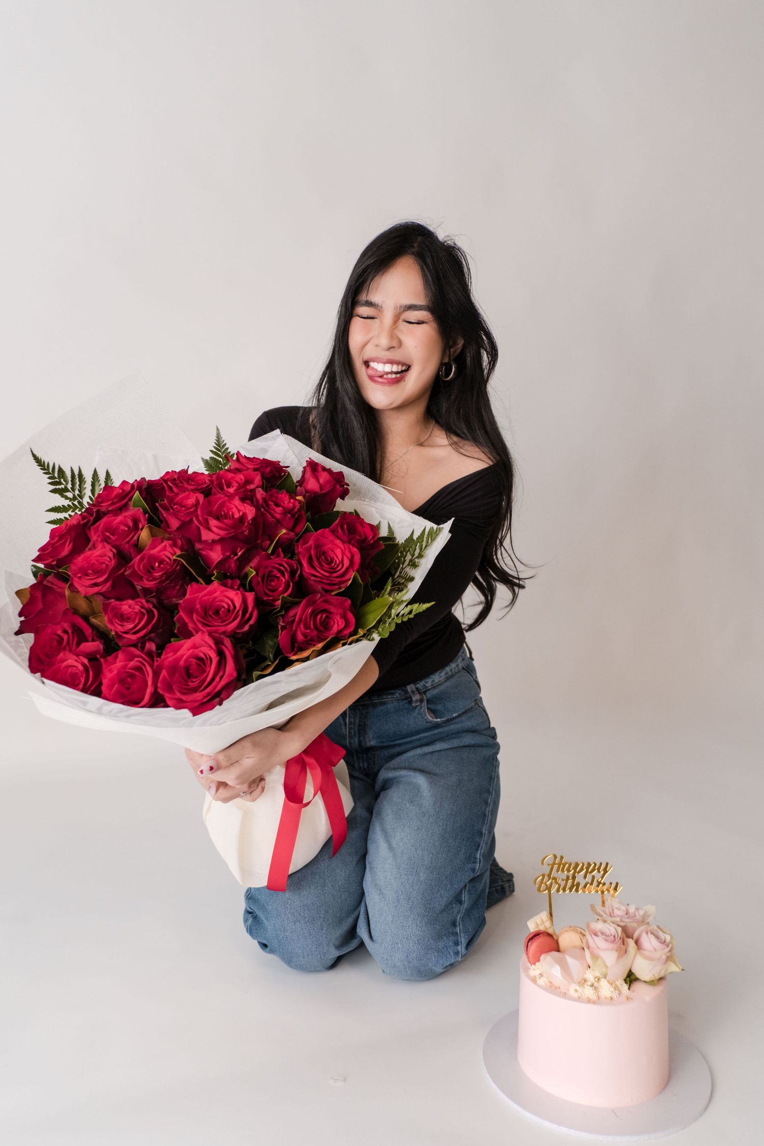 Young woman kneeling joyfully holding large red rose bouquet beside pink Happy Birthday cake on white background.