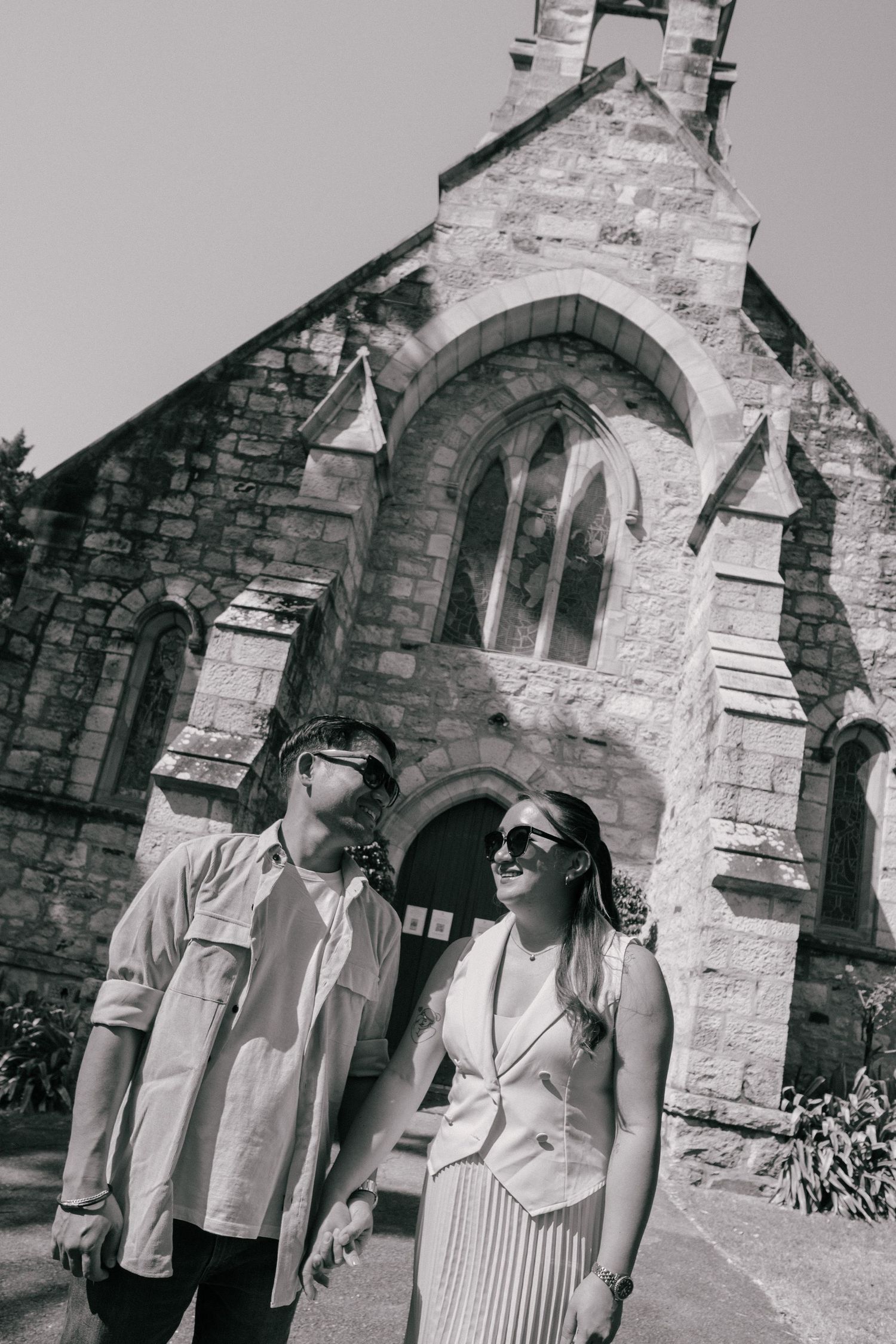 Black and white photo of a couple holding hands and smiling outside a historic stone church on a sunny day.