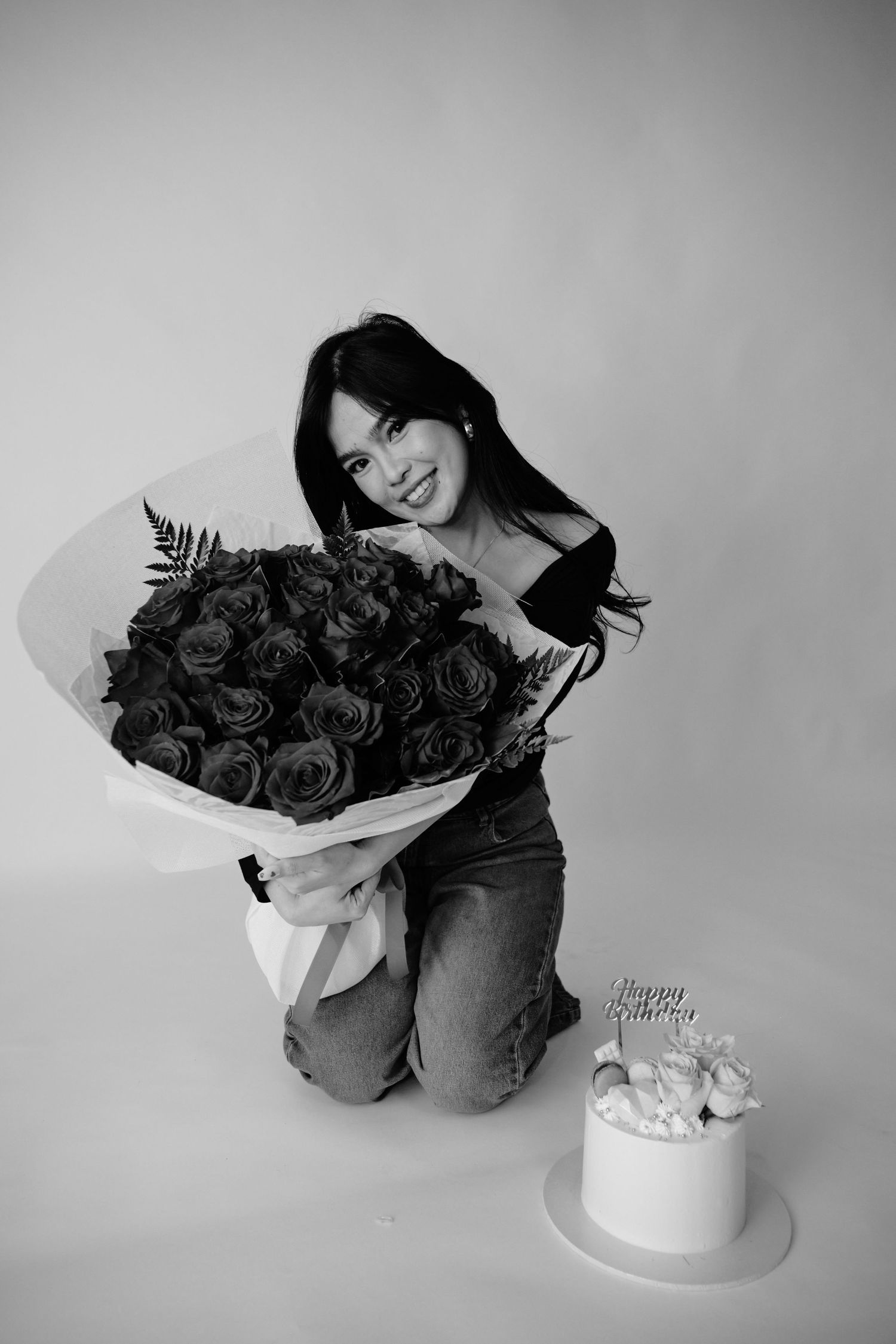 Young woman kneeling with large rose bouquet beside a Happy Birthday cake in black and white photo.