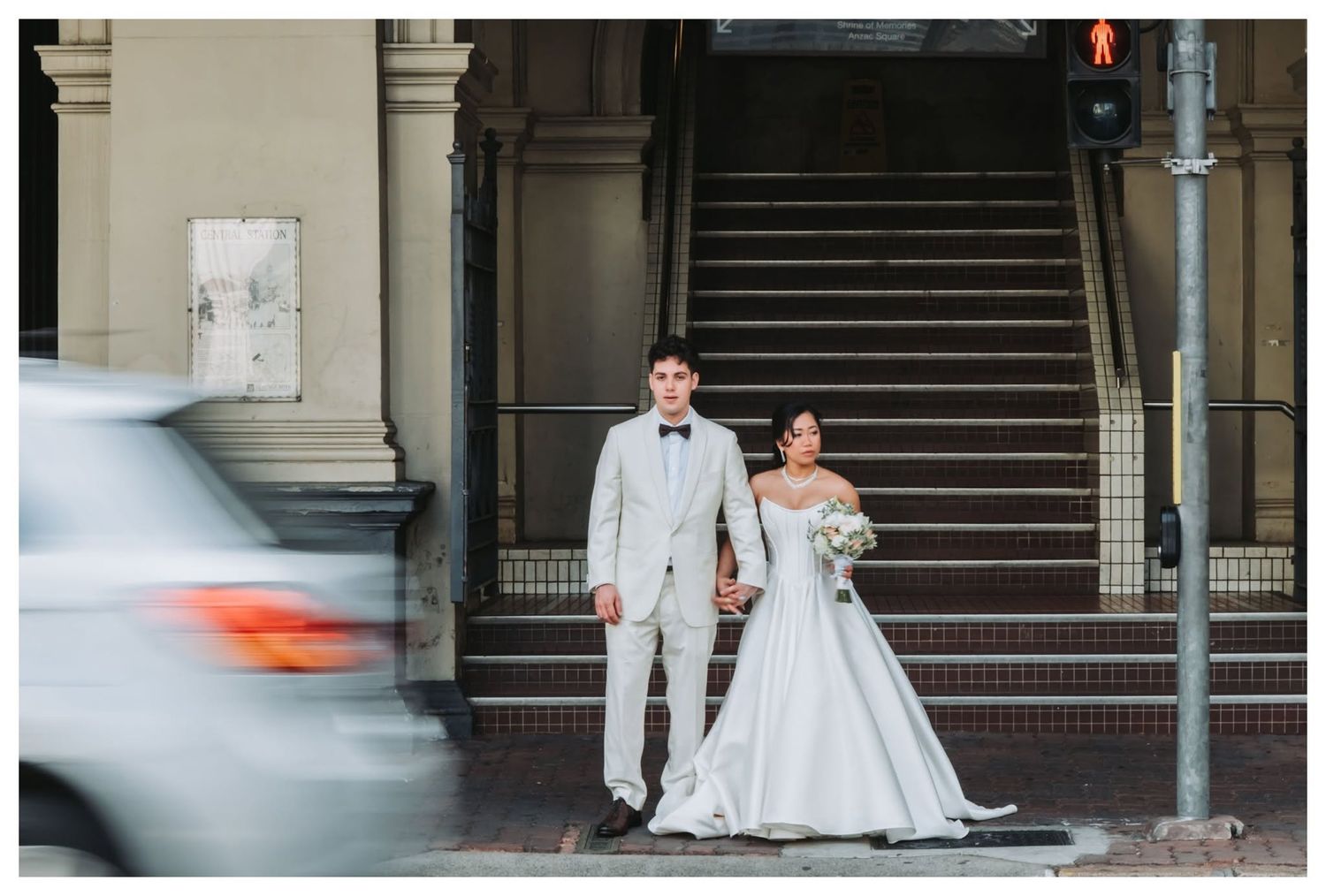 Bride and groom in elegant wedding attire standing at a city intersection with a car passing by.