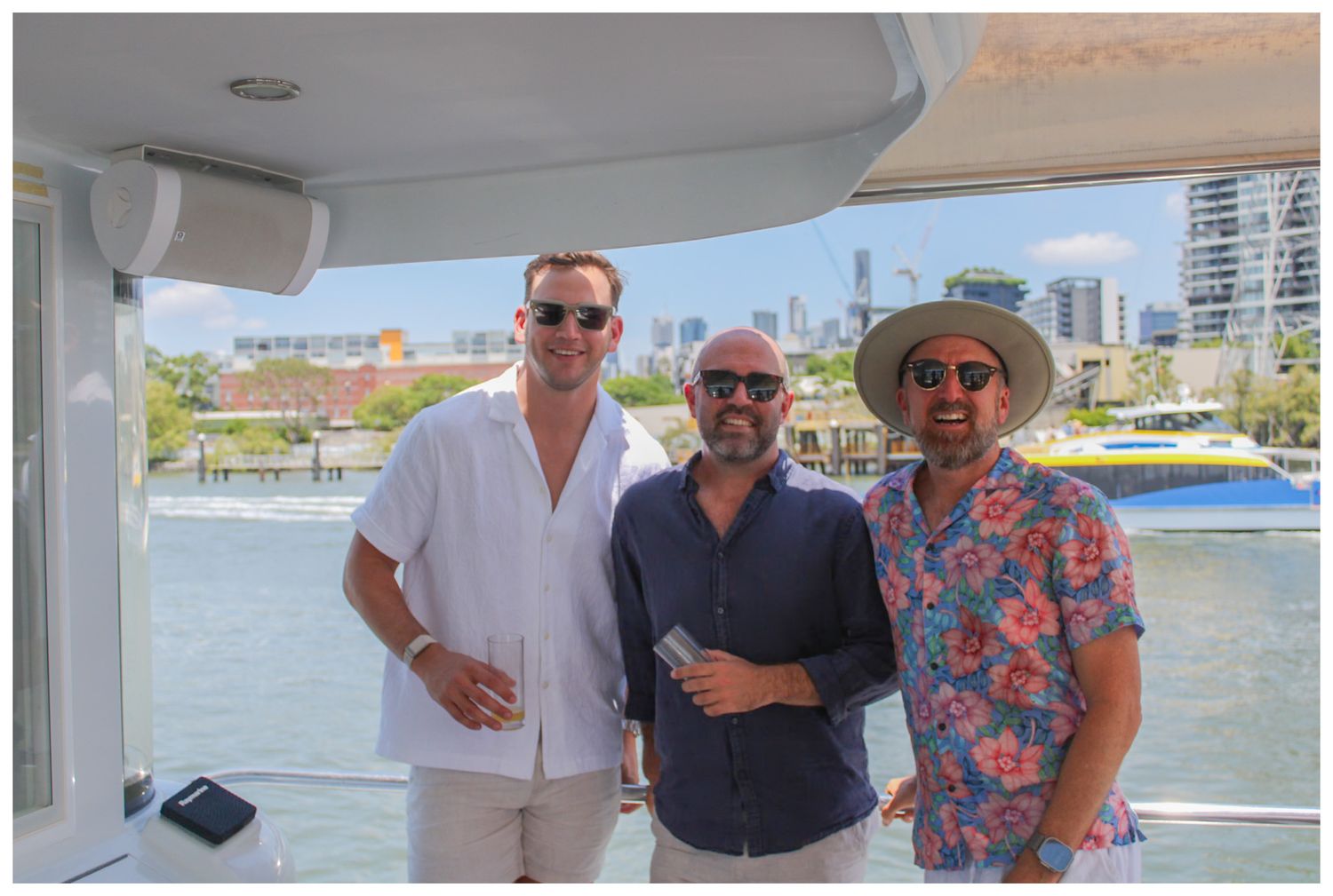 Three men smiling together on a boat with city skyline and waterway in the background.