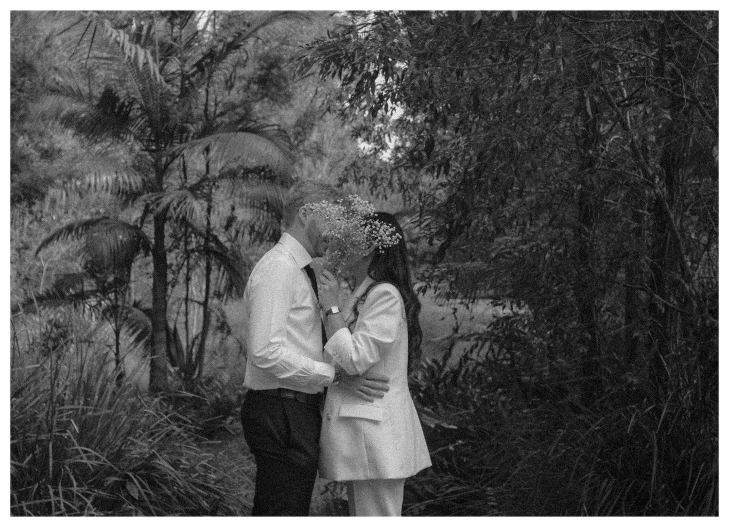 Black and white romantic photo of a couple embracing in a lush green forest setting.