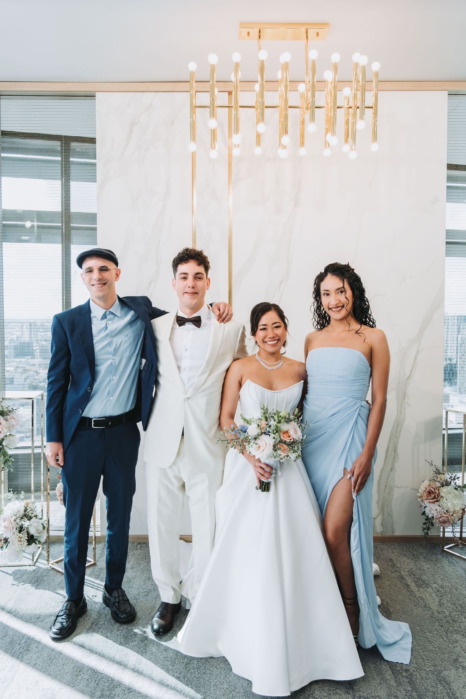 Wedding party poses under gold chandelier, bride in white gown with bouquet, groom in white suit, bridesmaid in blue dress.