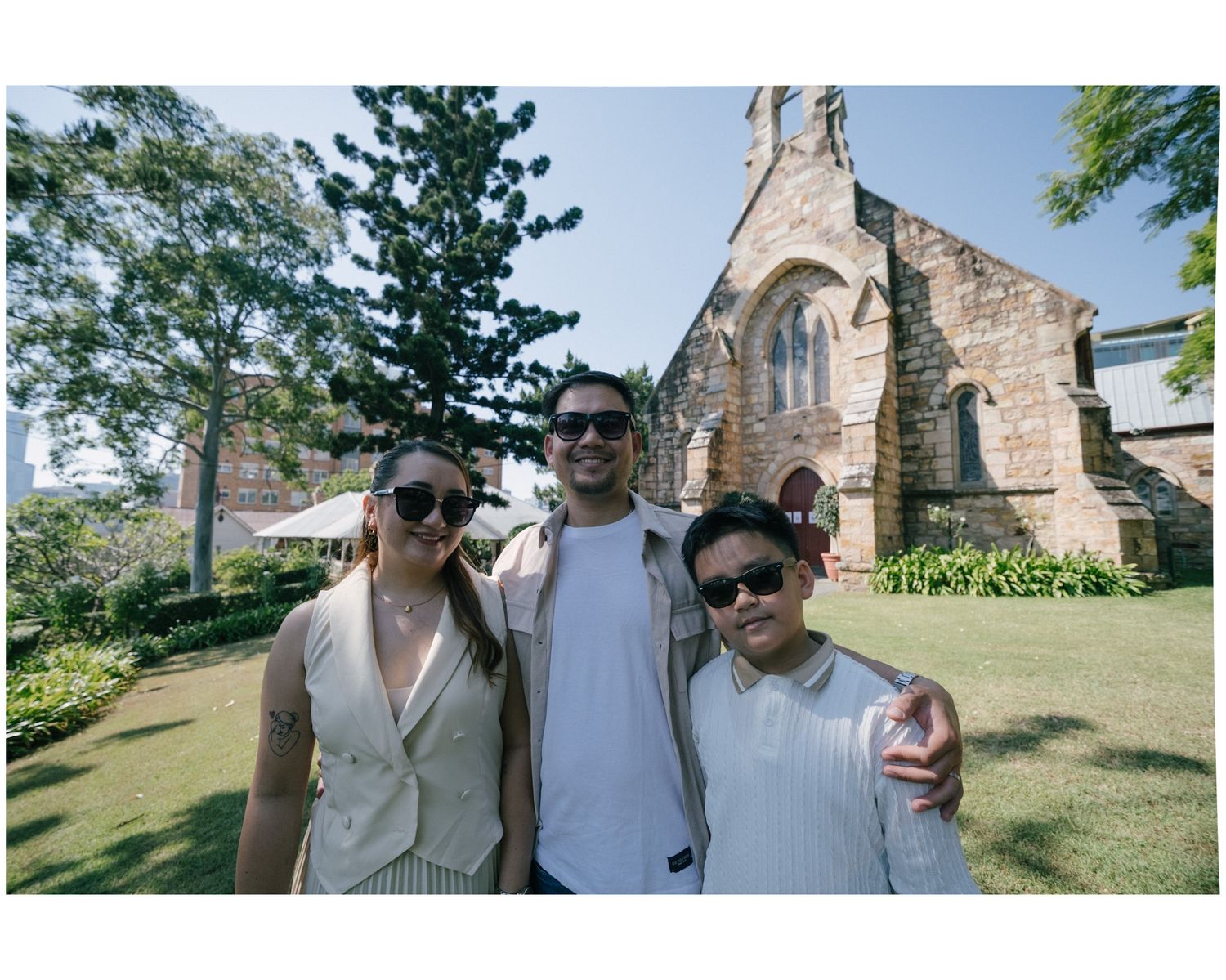 Three friends posing together in front of a historic stone church surrounded by lush green gardens.