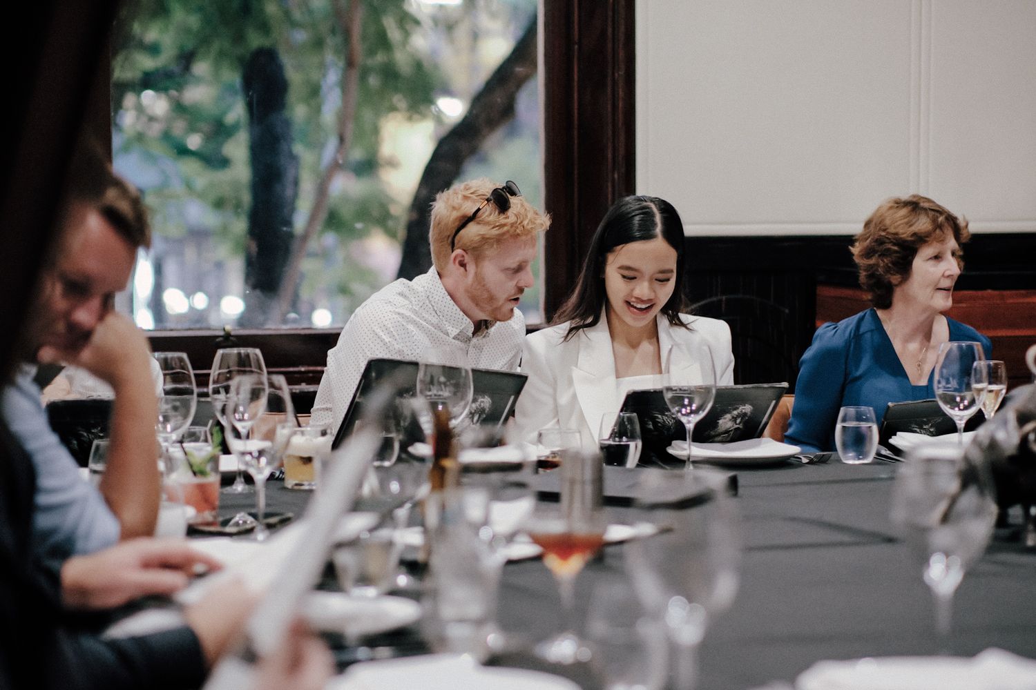Elegantly dressed guests laughing and socializing at a formal restaurant dining table with wine glasses.