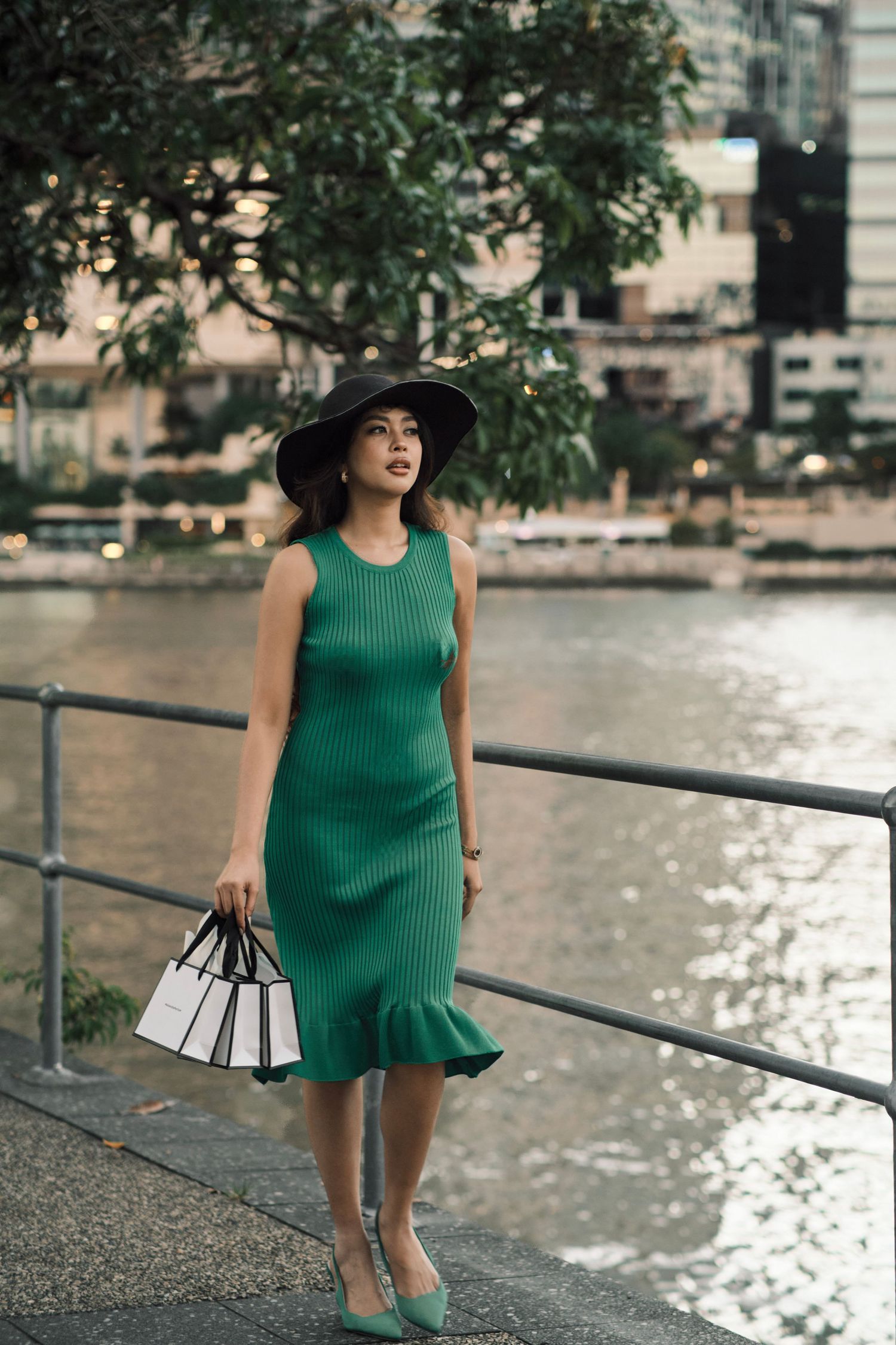Woman in green ribbed midi dress and black wide-brim hat holding shopping bags by a city waterfront.