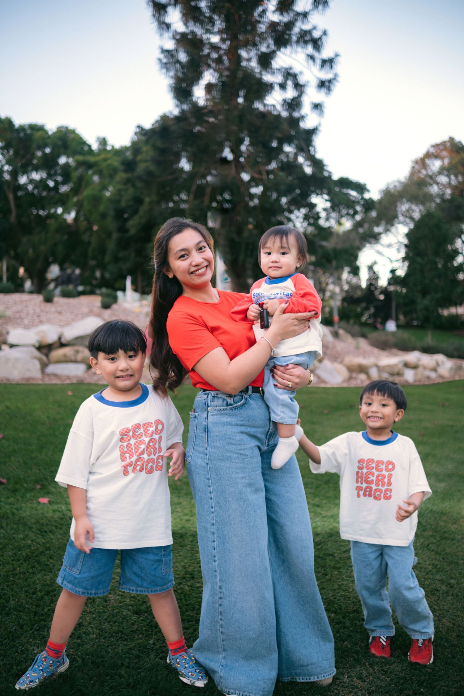 Asian mother in red top and jeans holds baby while two young boys in matching white shirts pose outdoors.