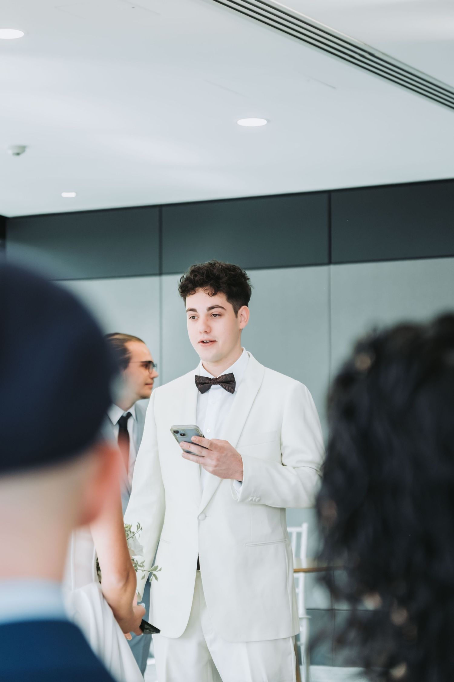 Young man in white tuxedo with bow tie holds phone at formal wedding event.