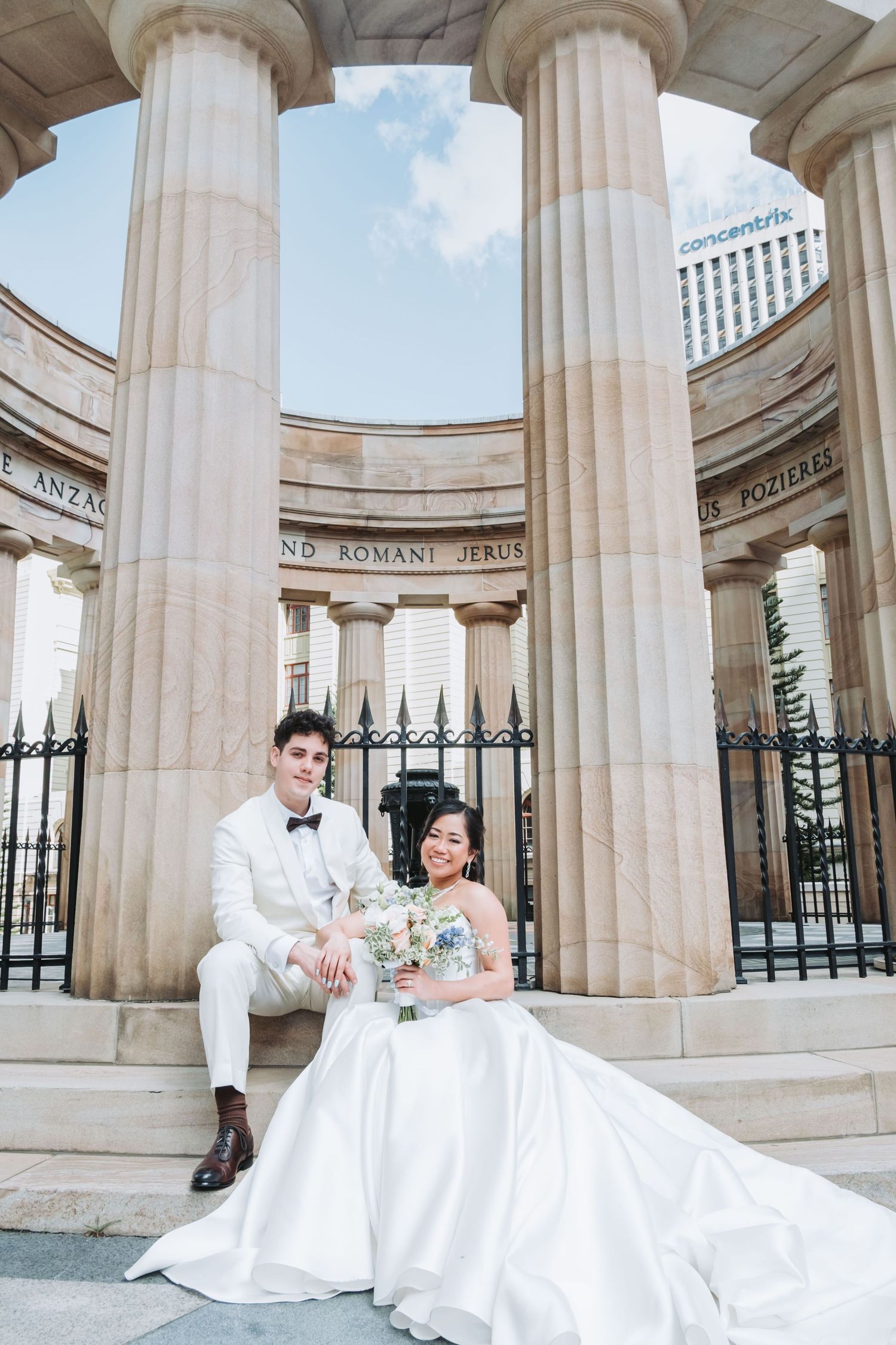 A bride and groom pose elegantly at a classical stone colonnade monument on their wedding day.