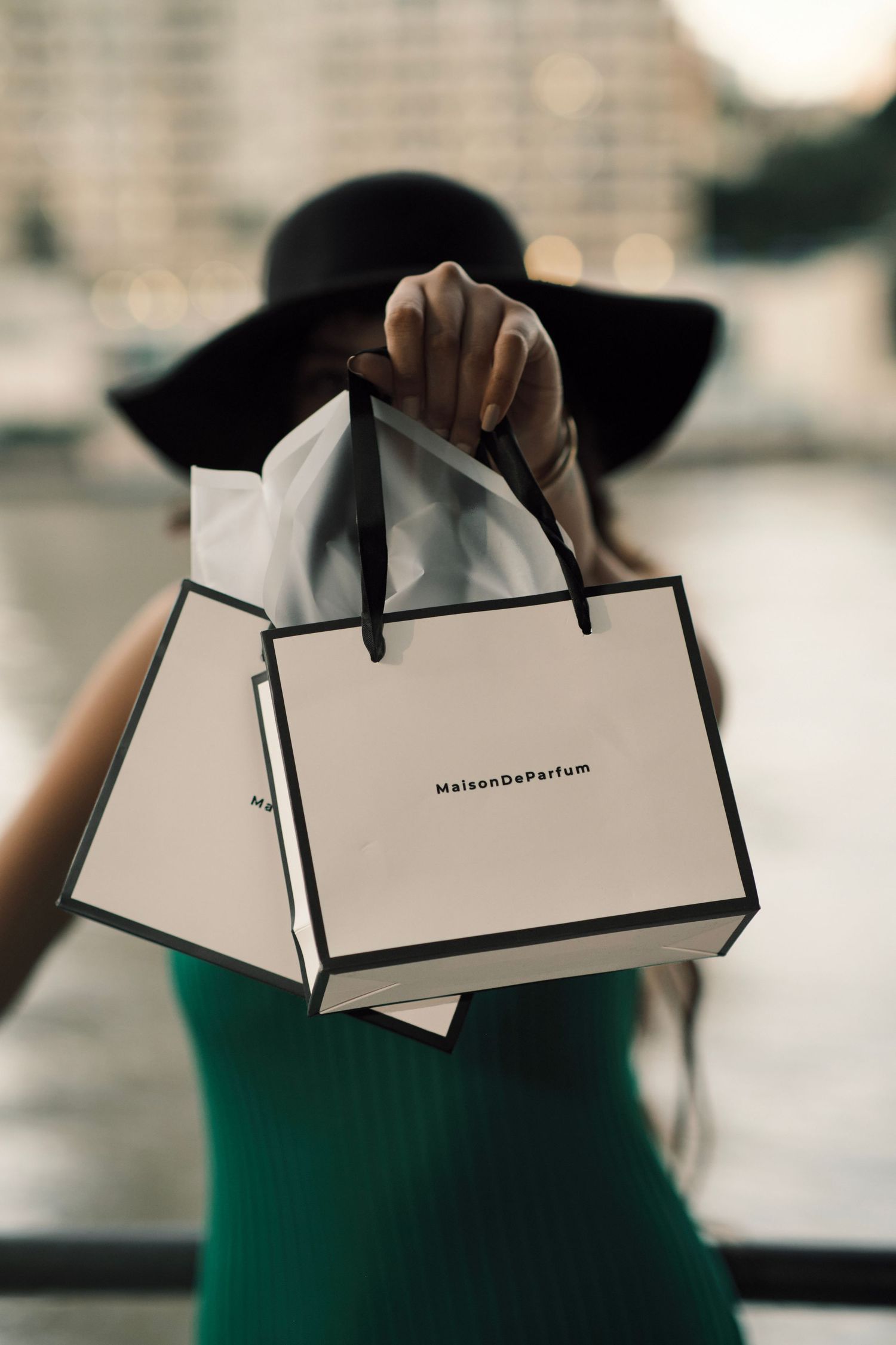 Woman in green dress and black hat holding a MaisonDeParfum luxury shopping bag outdoors.