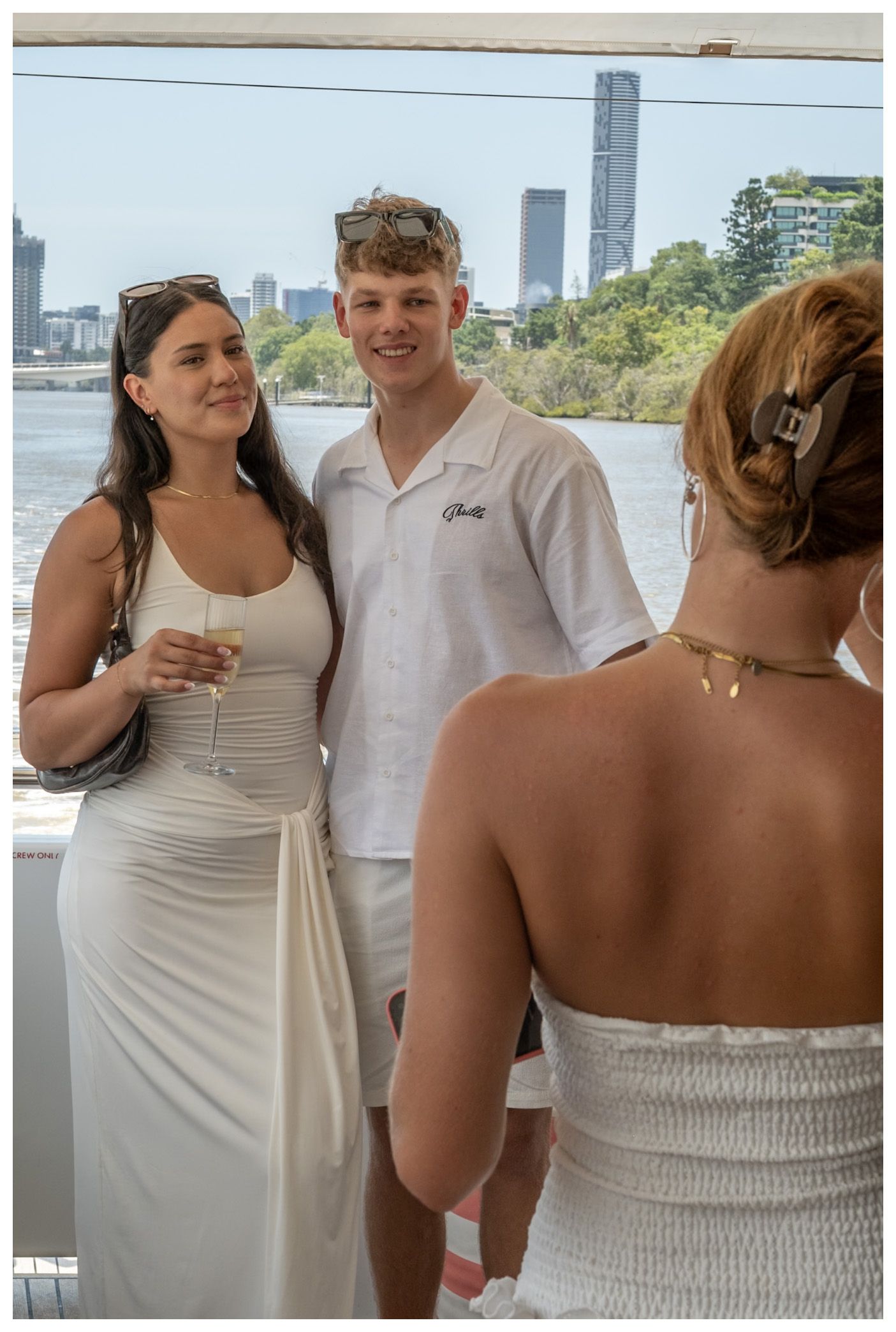 Three young adults in white outfits socializing on a boat with city skyline views on a sunny day.