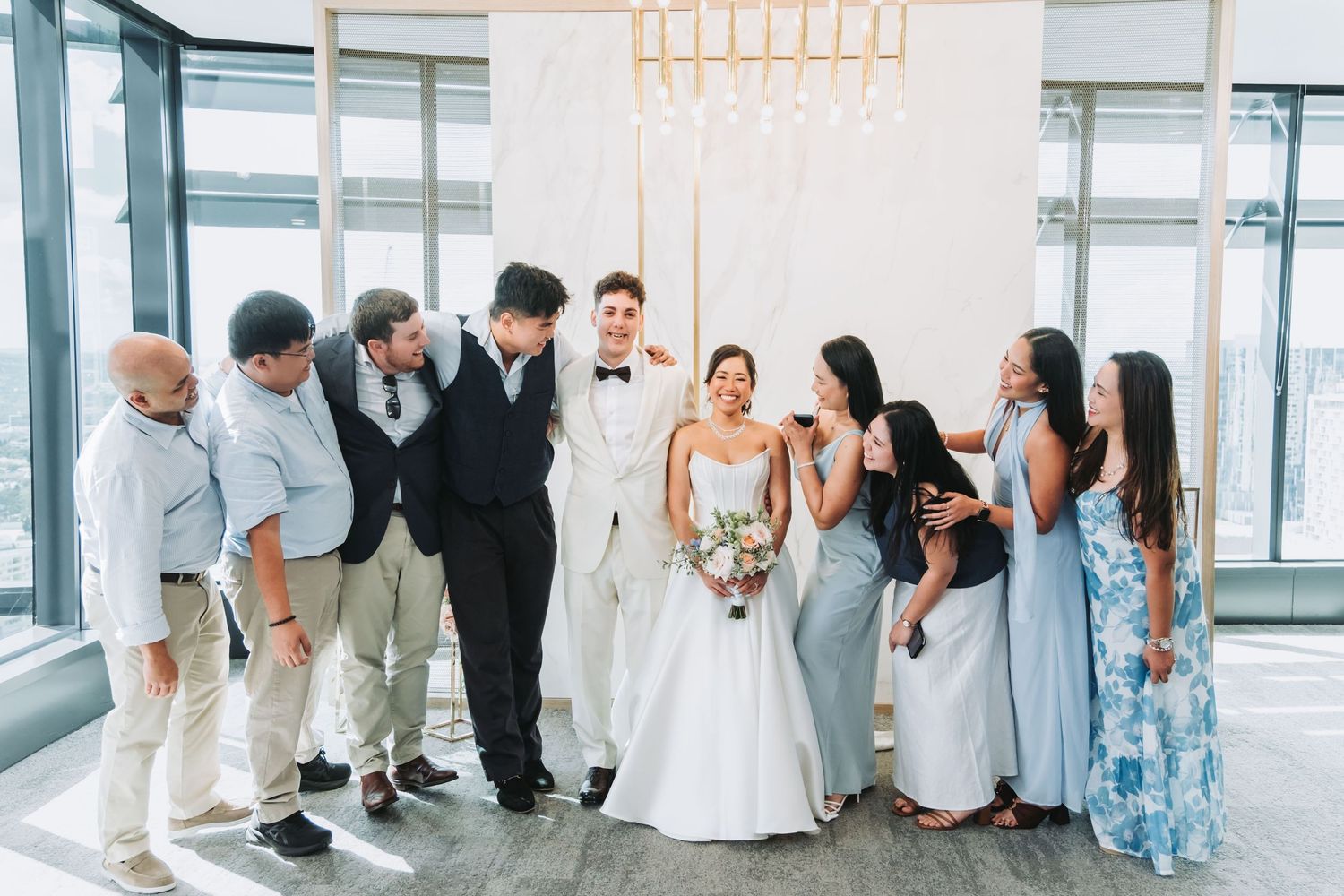 Wedding party gathers around bride and groom in elegant venue with floor-to-ceiling windows and gold chandelier backdrop.