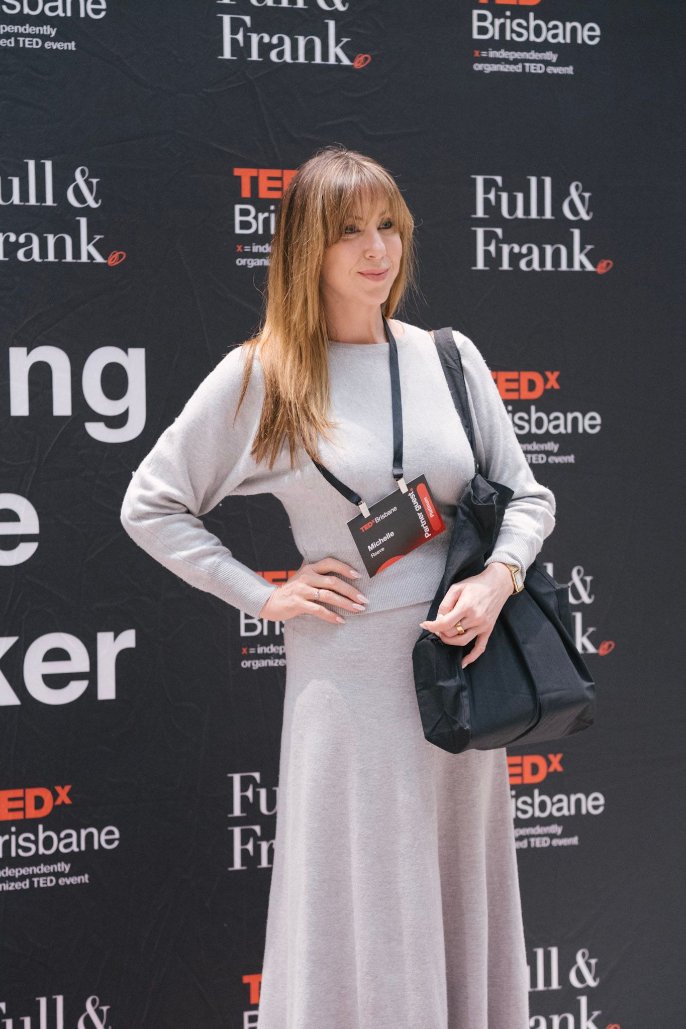 Woman in grey dress poses at TEDx Brisbane event backdrop sponsored by Full & Frank.