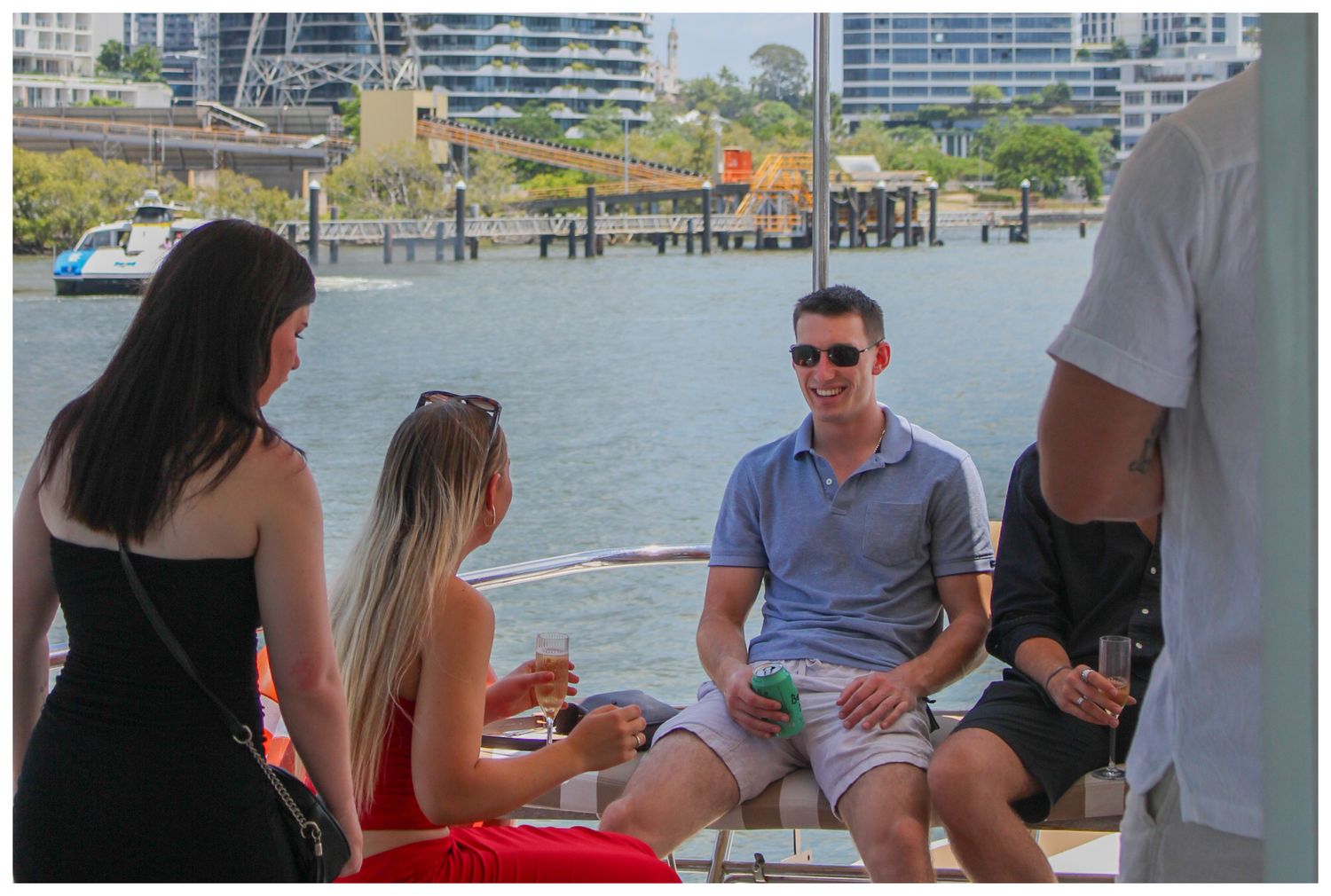 Group of people socializing on a boat deck with city buildings and waterway visible behind them.