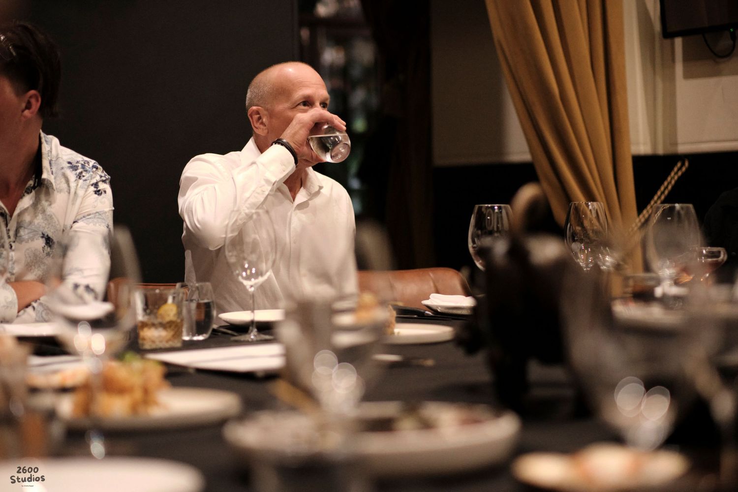 A candid shot of a man drinking at an event, with blurred guests in the foreground.