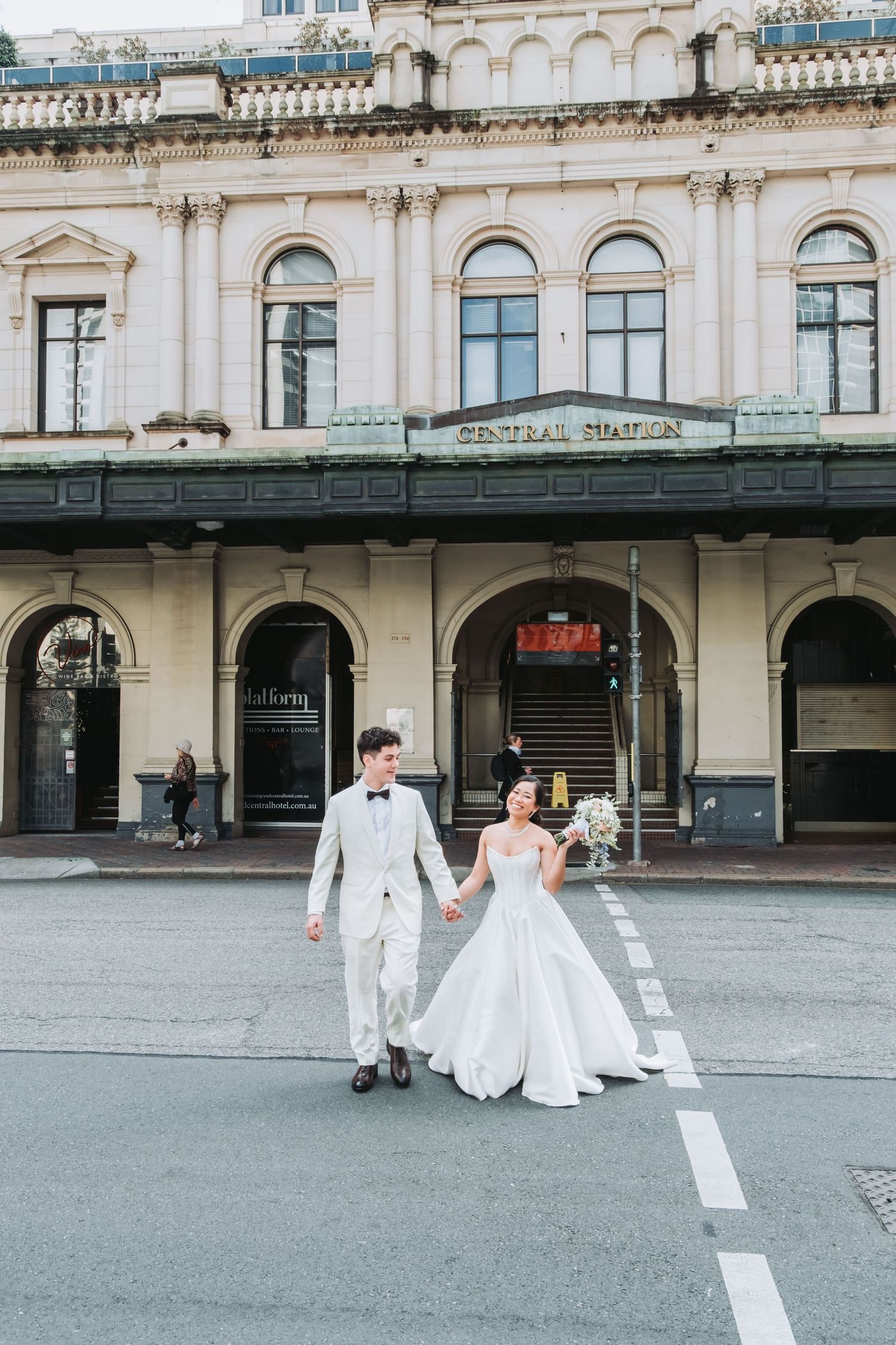 Bride and groom walk hand-in-hand across a street in front of Central Station in elegant wedding attire.