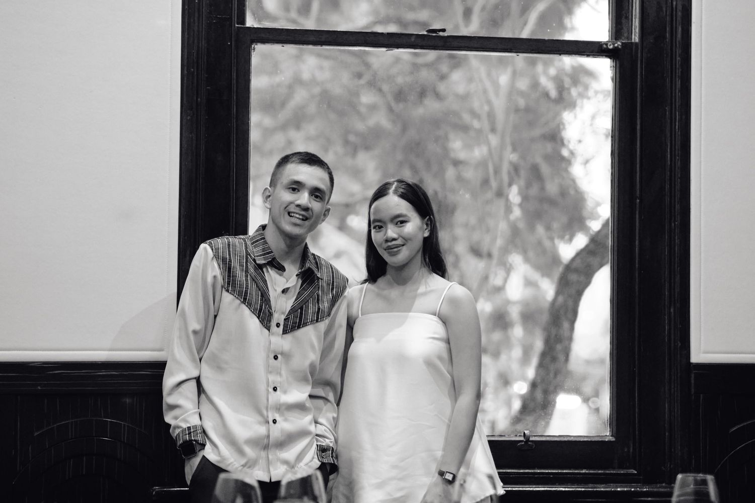 Black and white portrait of smiling couple standing together near large window indoors.