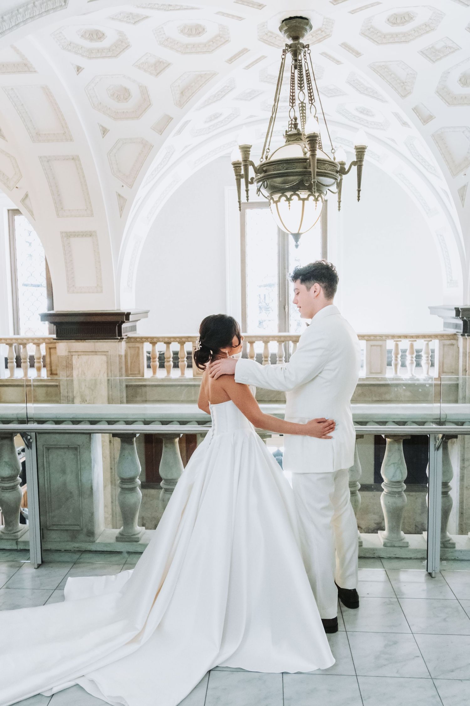 Bride and groom share a romantic moment in an elegant white ballroom with arched ceilings and a chandelier.
