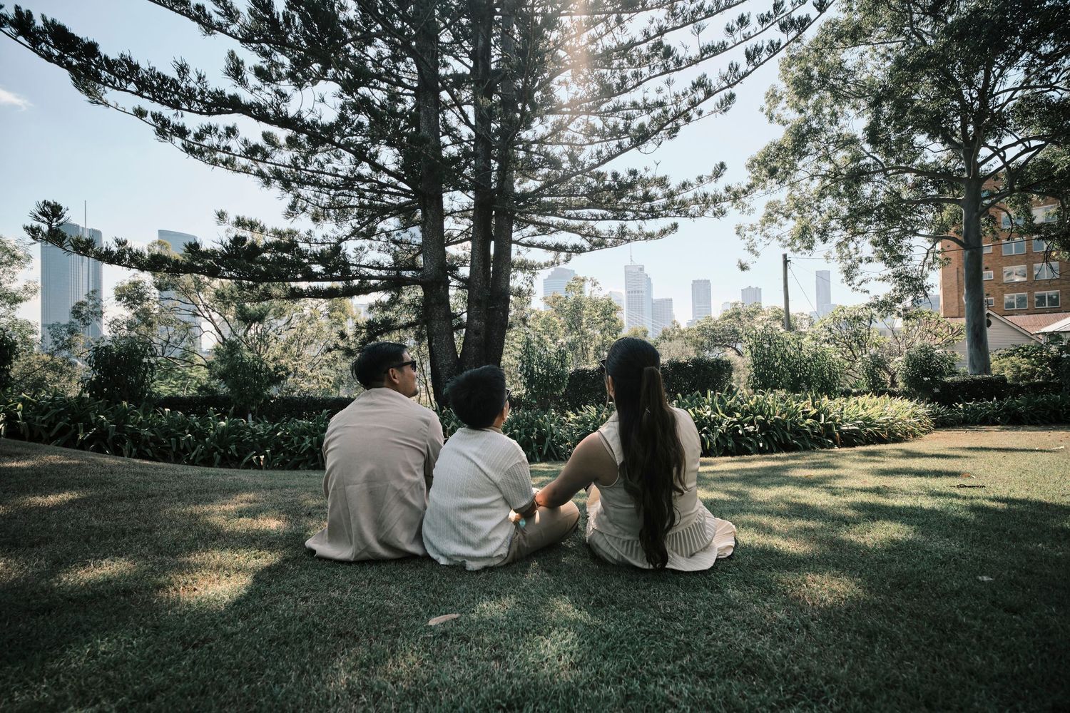 A family of three sitting on grass under a tree, viewed from behind.