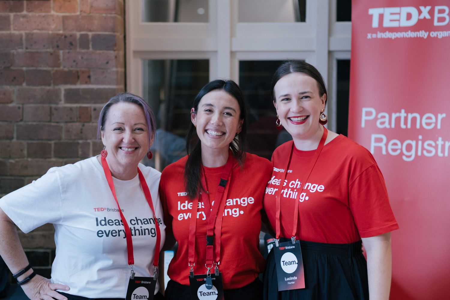 Three women in red TEDx t-shirts smile together at a TEDx event, with a TEDx banner visible behind them.
