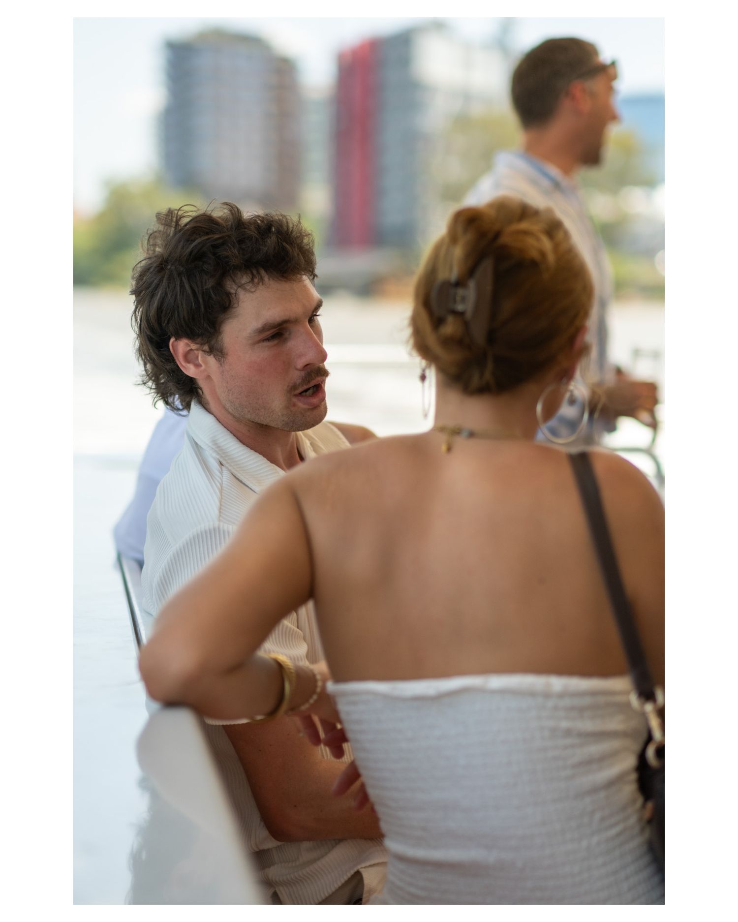 Two people in white conversing outdoors near water, with a city skyline visible in the background.