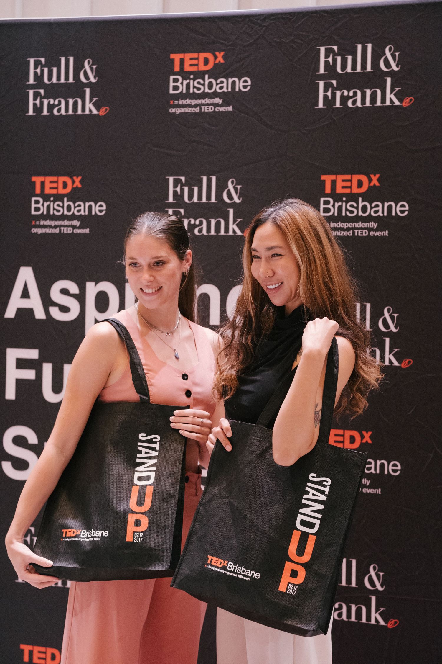 Two women smile holding TEDx Brisbane Stand UP tote bags at a Full & Frank sponsored event backdrop.