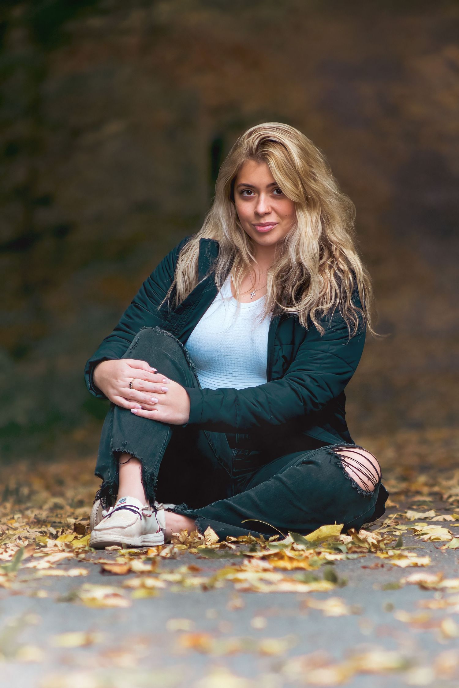Blonde woman sitting cross-legged on ground surrounded by autumn leaves, wearing ripped jeans and dark blazer.
